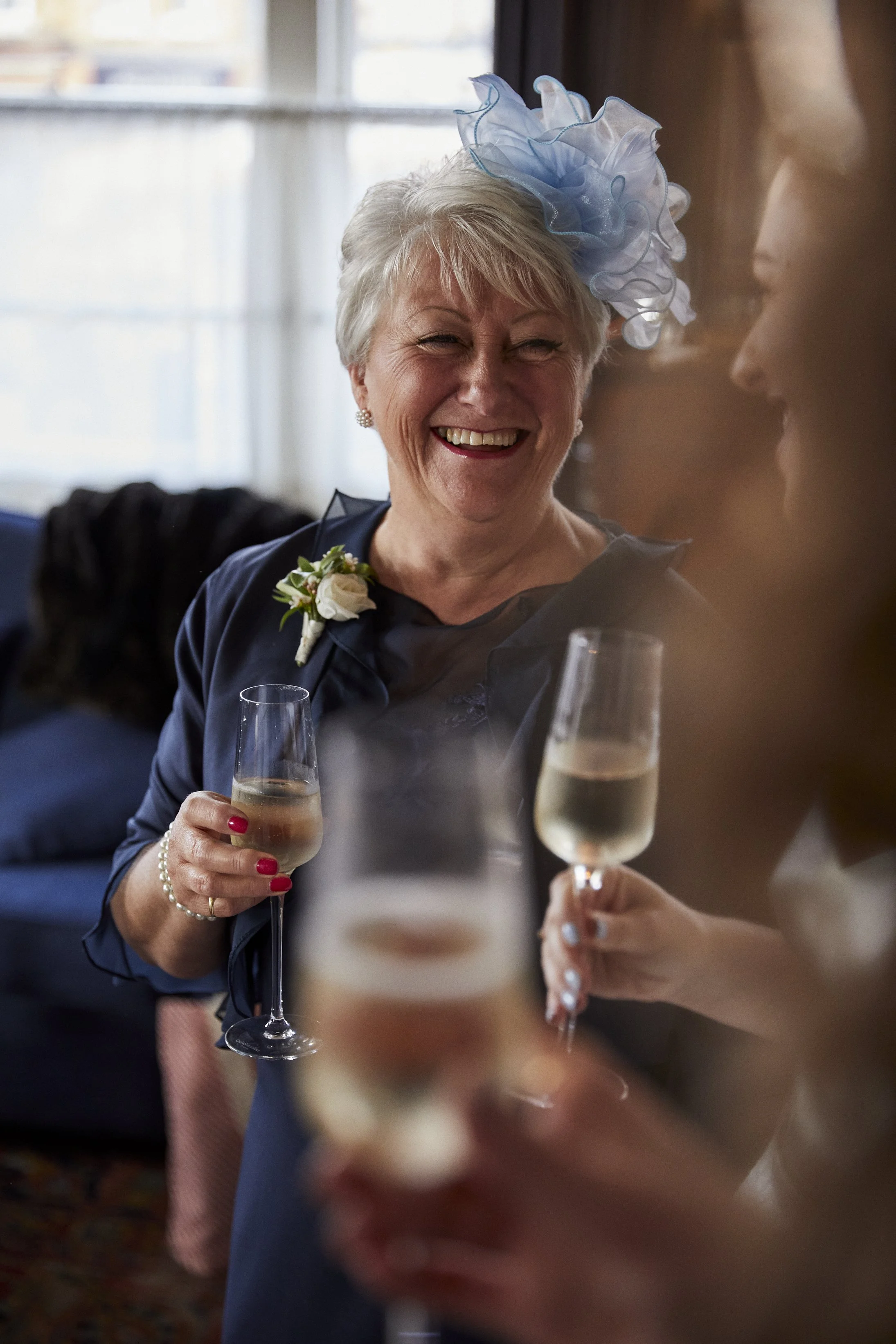 An older woman with short gray hair wearing a blue fascinator, smiling and holding a glass of champagne, part of a celebration with others with glasses of bubbly.