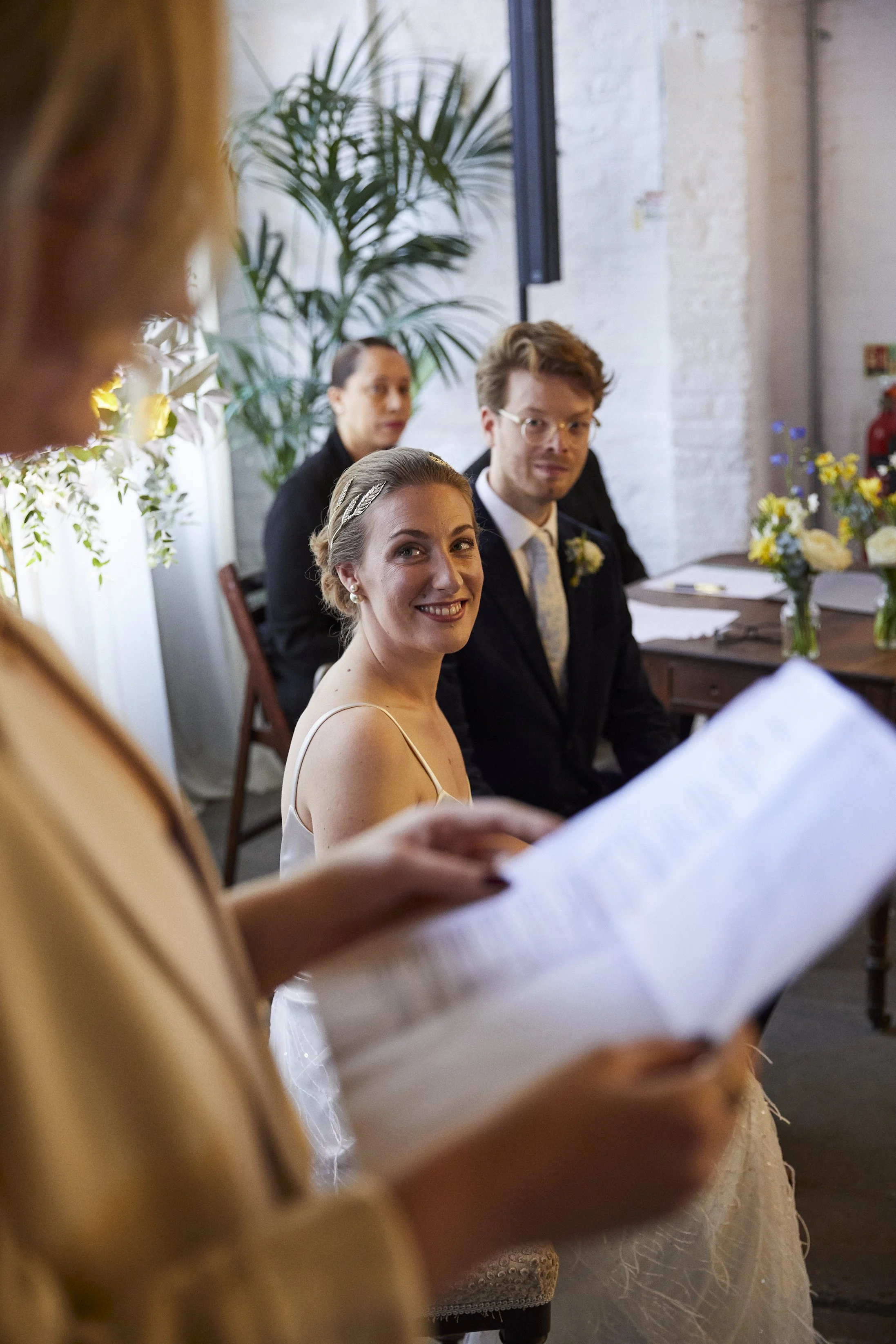 A bride and groom sitting next to each other, with two guests behind them, during a wedding ceremony. A woman in the foreground is reading from a paper.