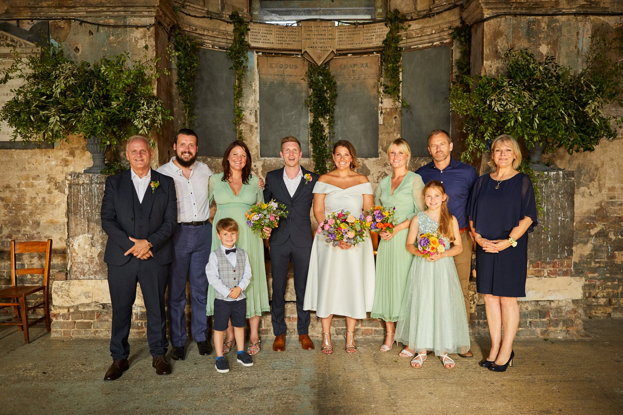 A group of people, including a bride and groom, posing for a wedding photo in front of an aged, rustic backdrop with greenery and old wooden chairs.