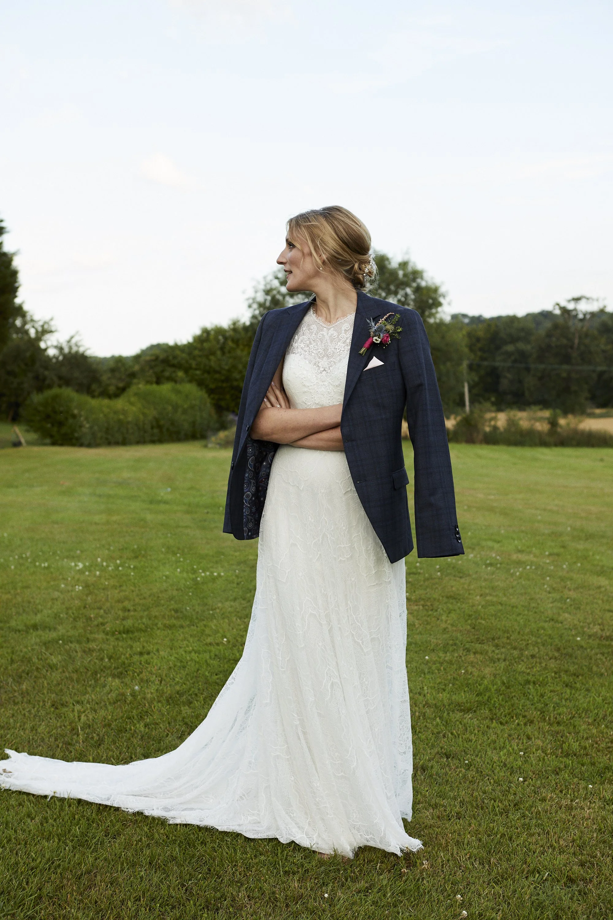 A woman in a white wedding dress with lace details and a navy blazer, standing outdoors on a grassy area with trees in the background, arms crossed, looking to her side.