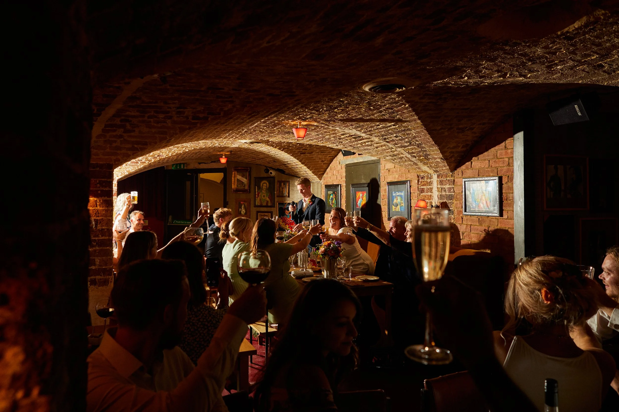 People at a wedding celebration or dinner party raising glasses for a toast inside a cozy brick-walled restaurant or wine cellar.