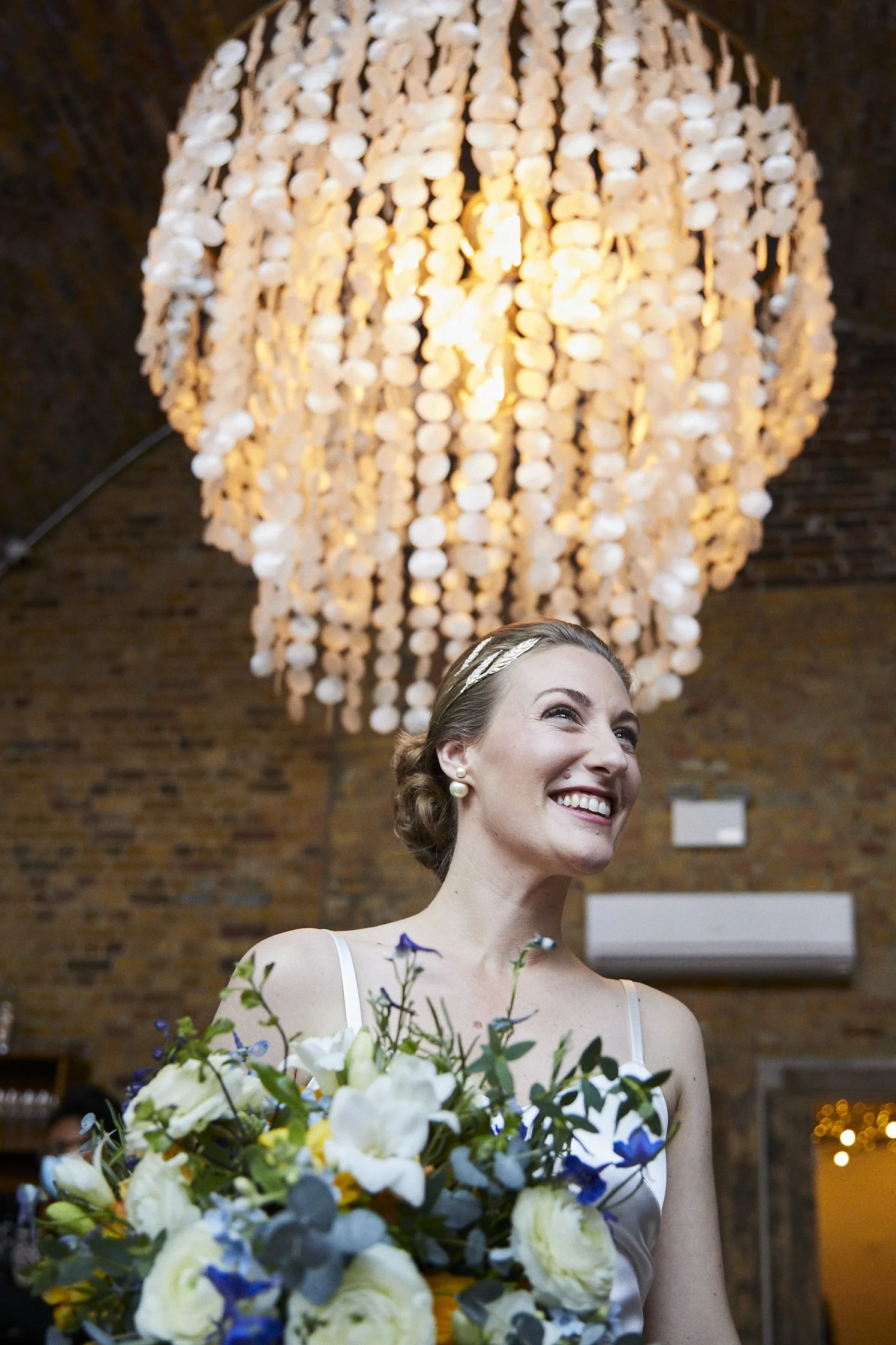 A woman smiling and holding a bouquet of flowers, standing in front of a large chandelier in what appears to be a brick-walled venue.