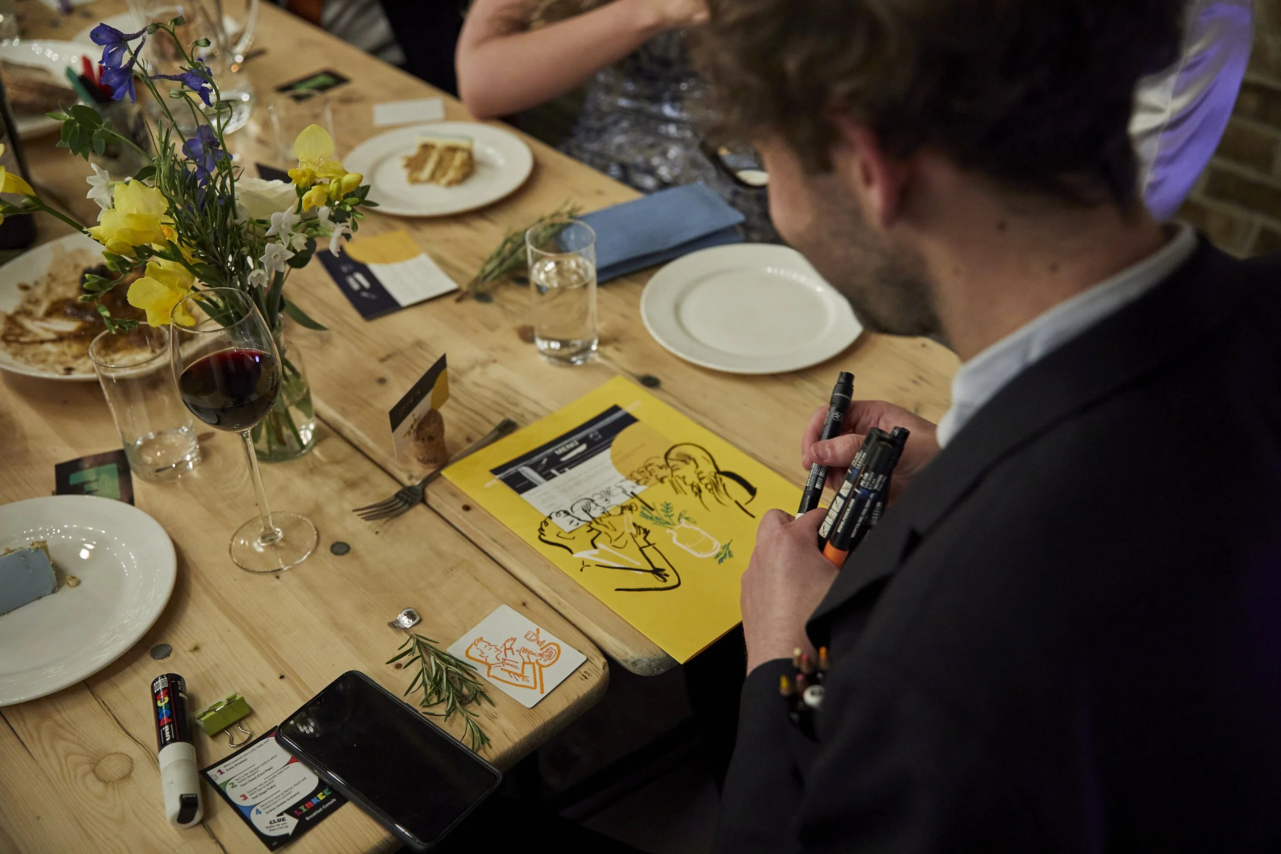 Man signing a yellow menu at a dining table with wine glasses, plates, flowers, and various items