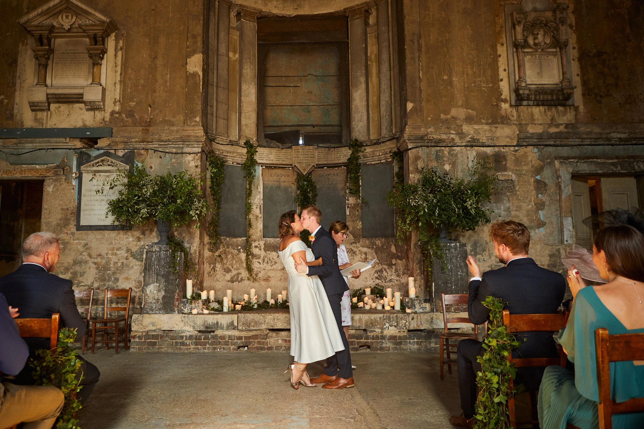 Couple sharing a kiss during their wedding ceremony inside a rustic, historic building, with guests seated on either side, surrounded by candles and greenery.