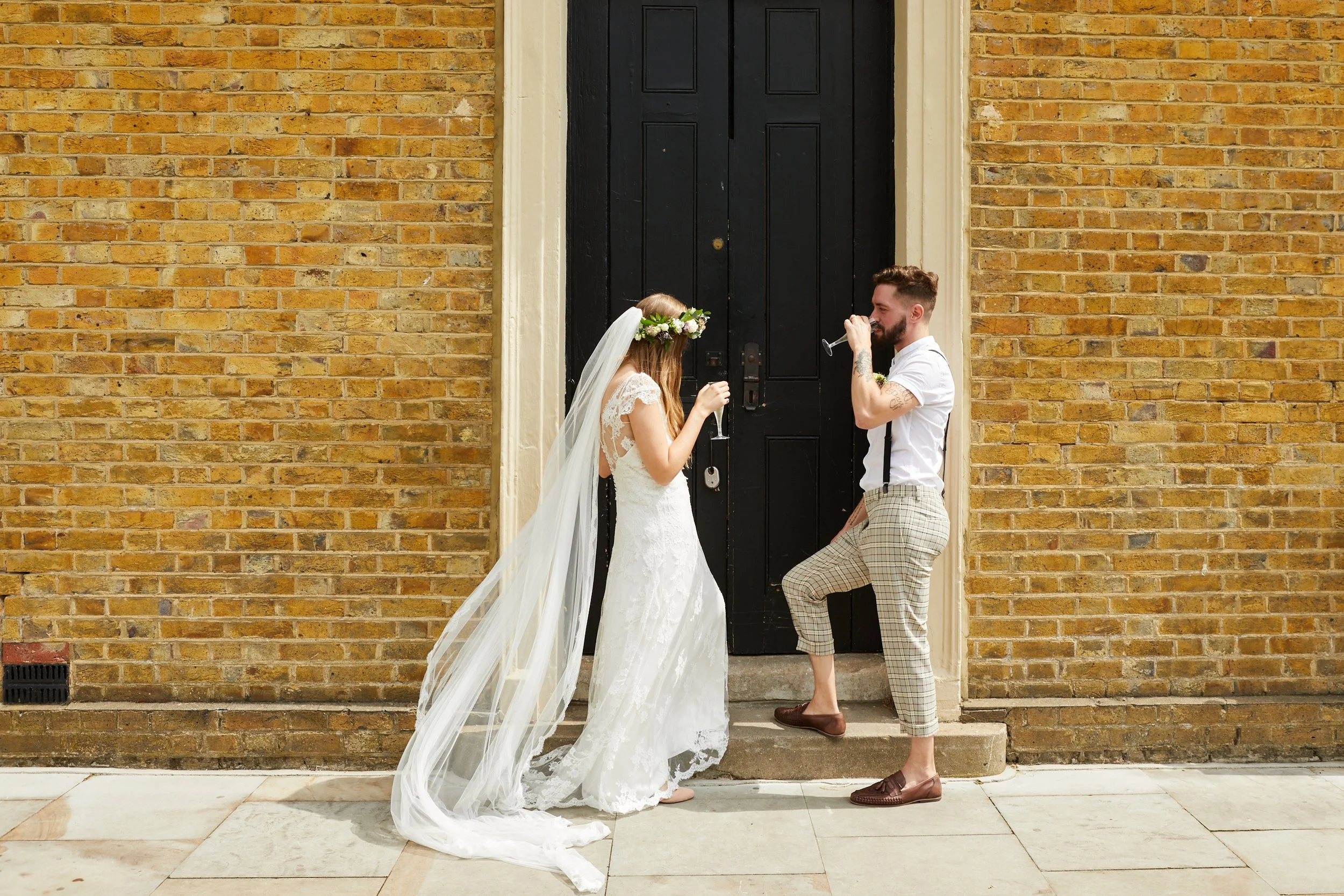 A bride and groom stand outside a brick building, each holding a champagne flute, sharing a toast in front of a black door. The bride wears a white lace wedding dress and a floral crown with a long veil. The groom wears a white shirt, plaid pants, and brown loafers.