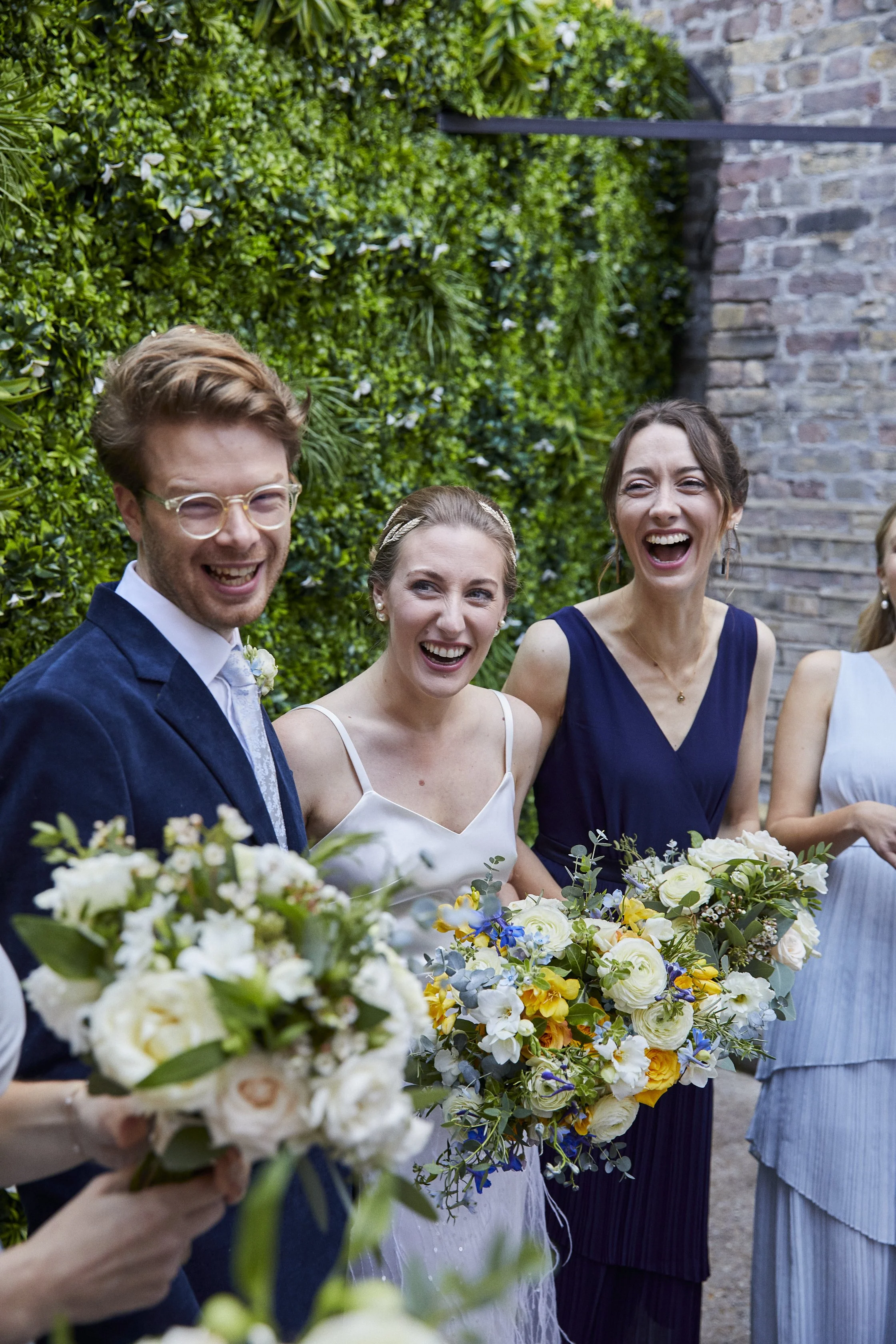 A group of people, including a man in glasses and a woman in a white dress, holding bouquets, are gathered outdoors, smiling and laughing at a wedding celebration.