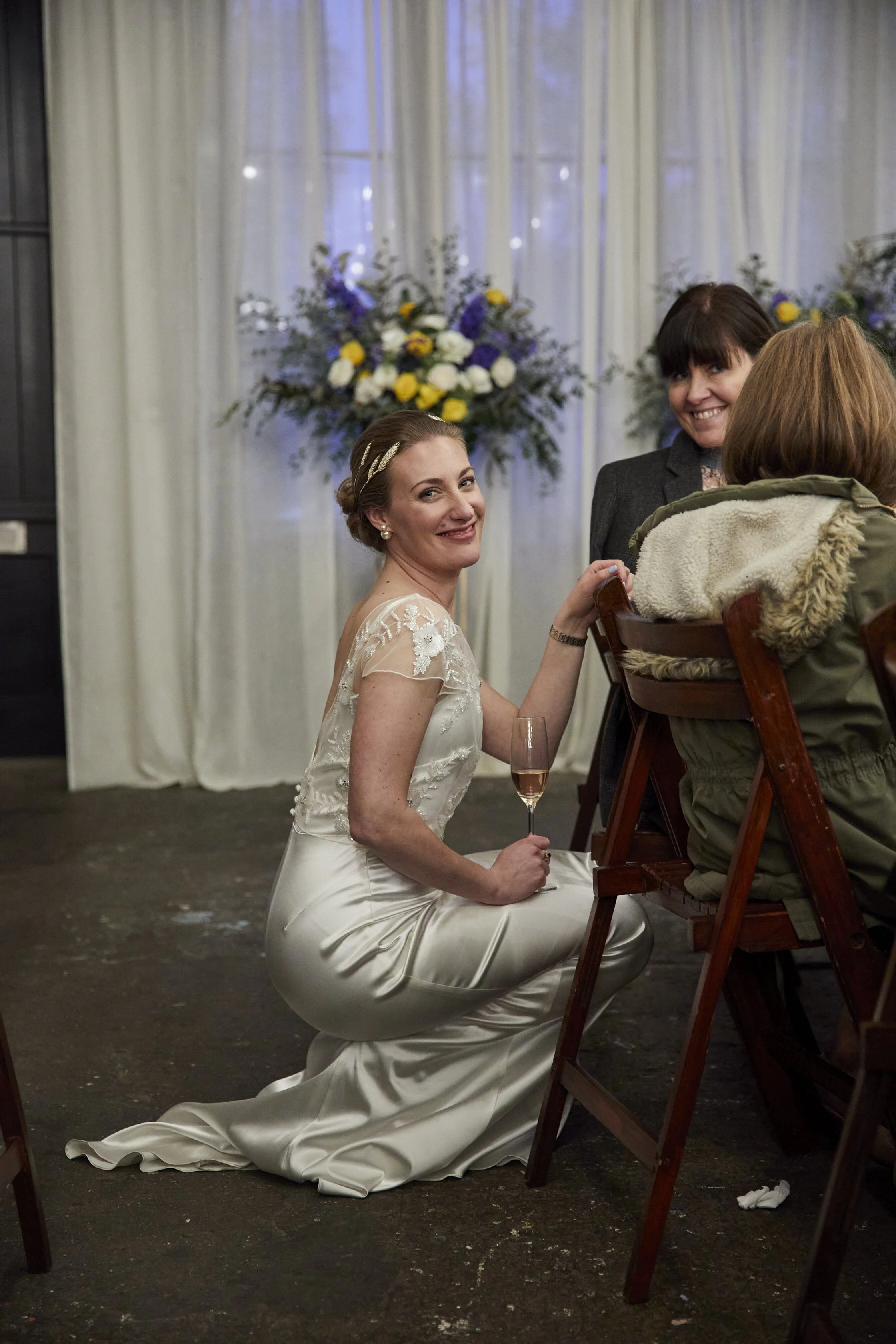 A bride kneeling on the floor in her wedding gown, smiling and holding a champagne glass, during a celebration with guests at a wedding reception.