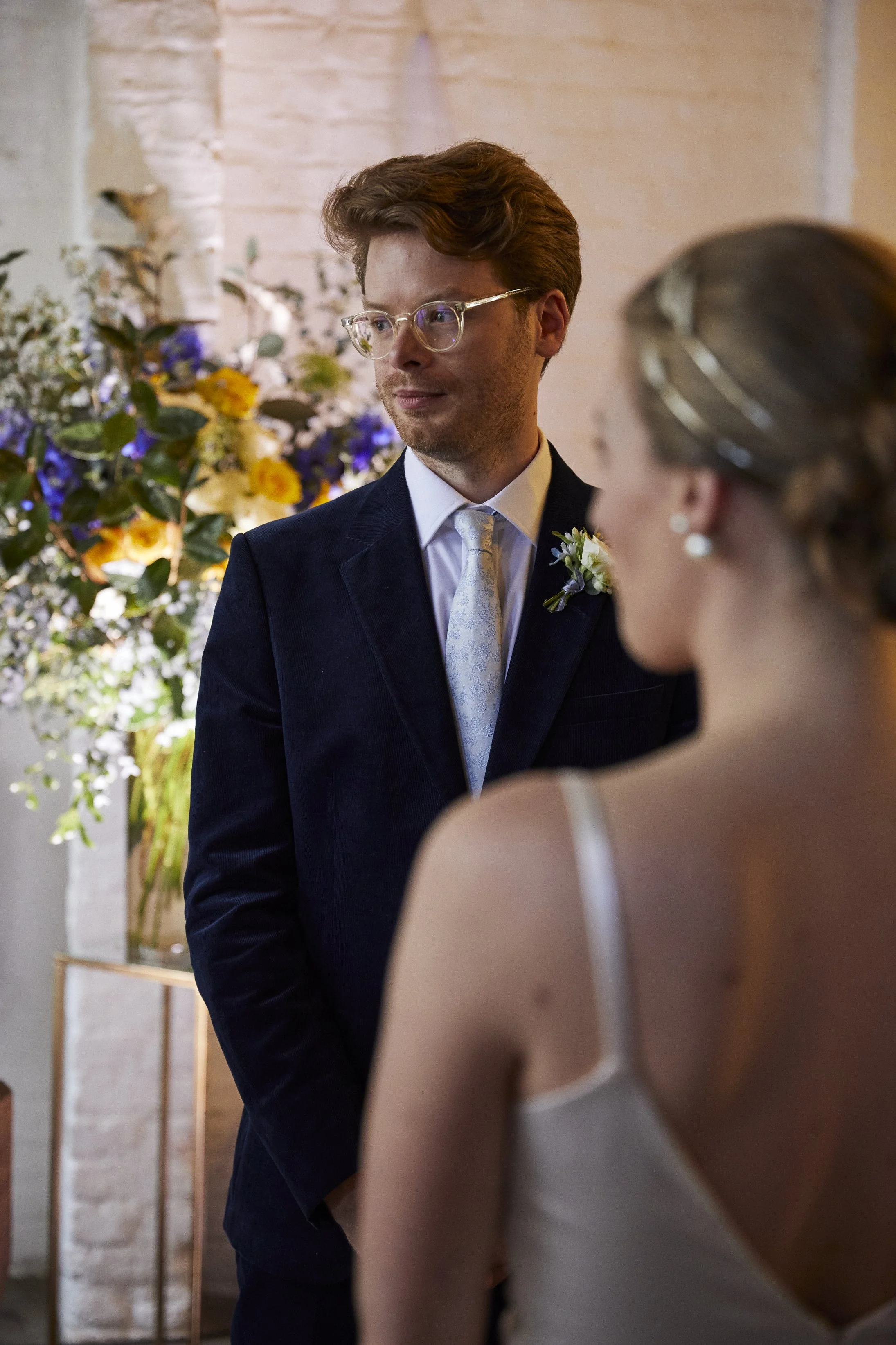 A groom wearing glasses, a dark suit, and a white tie stands during a wedding ceremony, looking at the bride. The bride is in a white dress with spaghetti straps, with her back to the camera, and her hair styled with a hair accessory. A floral arrang