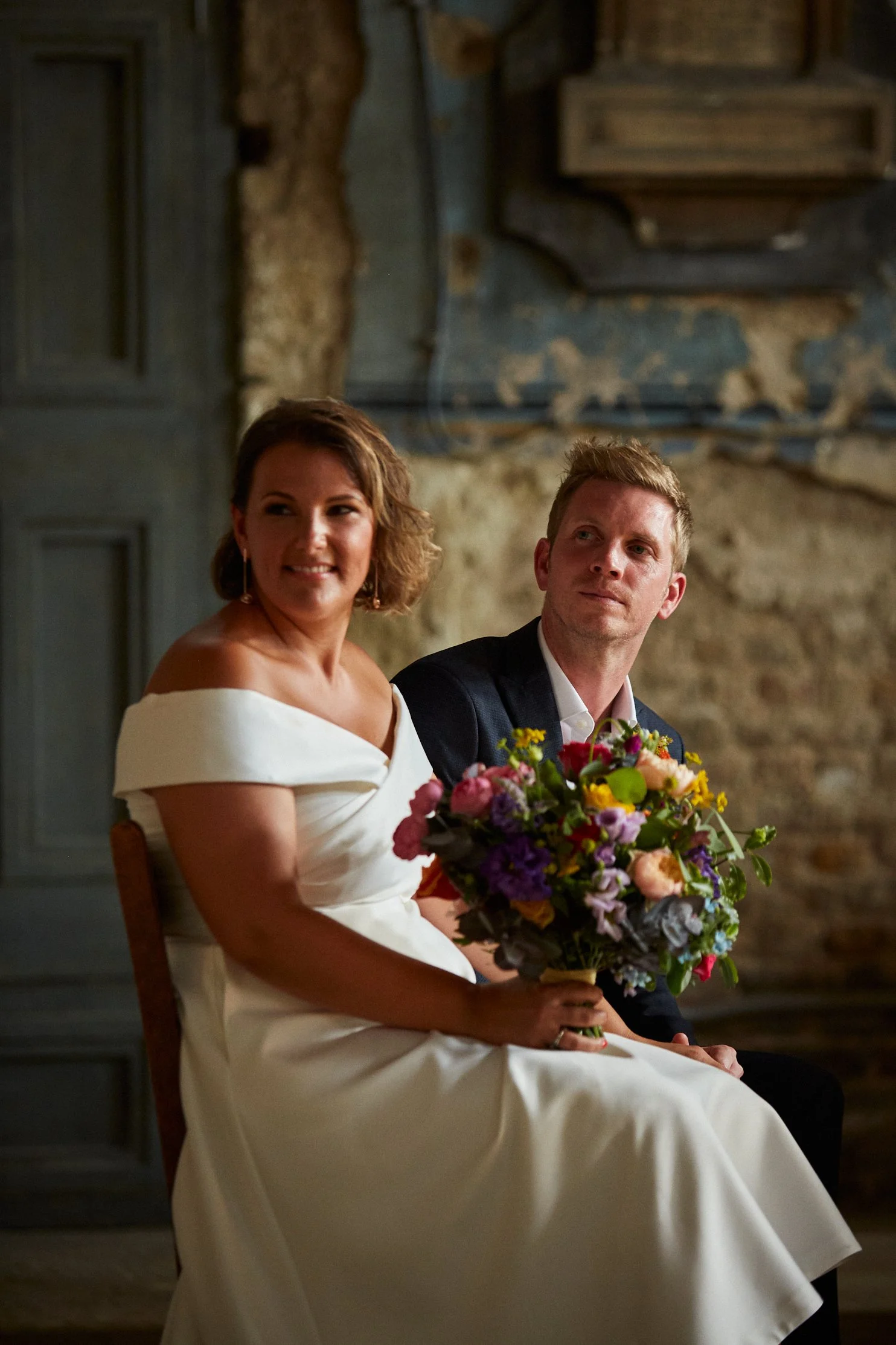 A bride in a white dress holding a colorful bouquet sits beside a groom in a dark suit, both sitting on chairs in a rustic setting with a textured stone wall.