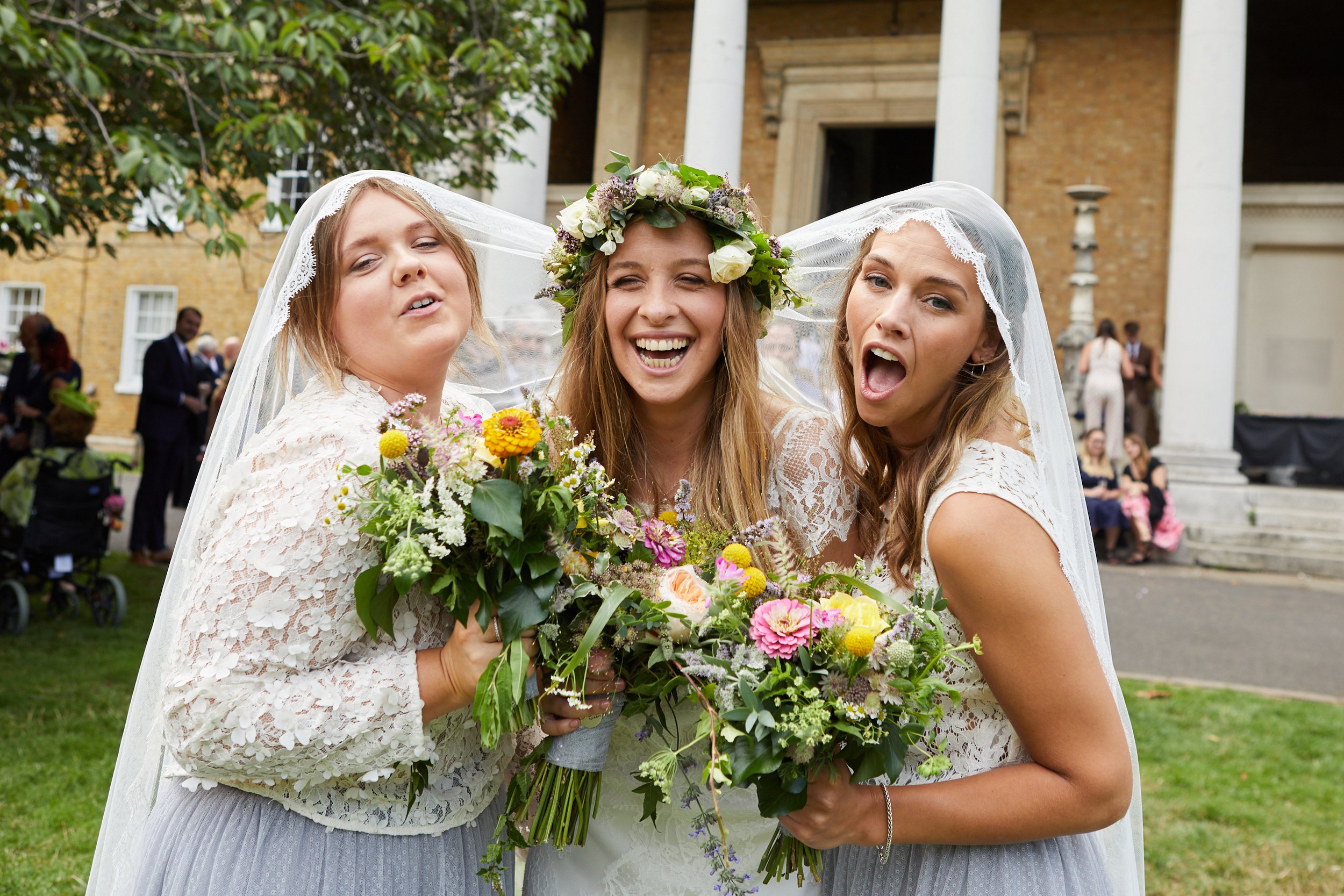 Three women dressed as bride and bridesmaids, smiling and posing for a photo outdoors on a wedding day, each holding a bouquet of colorful flowers, with a bride wearing a floral crown and a veil.