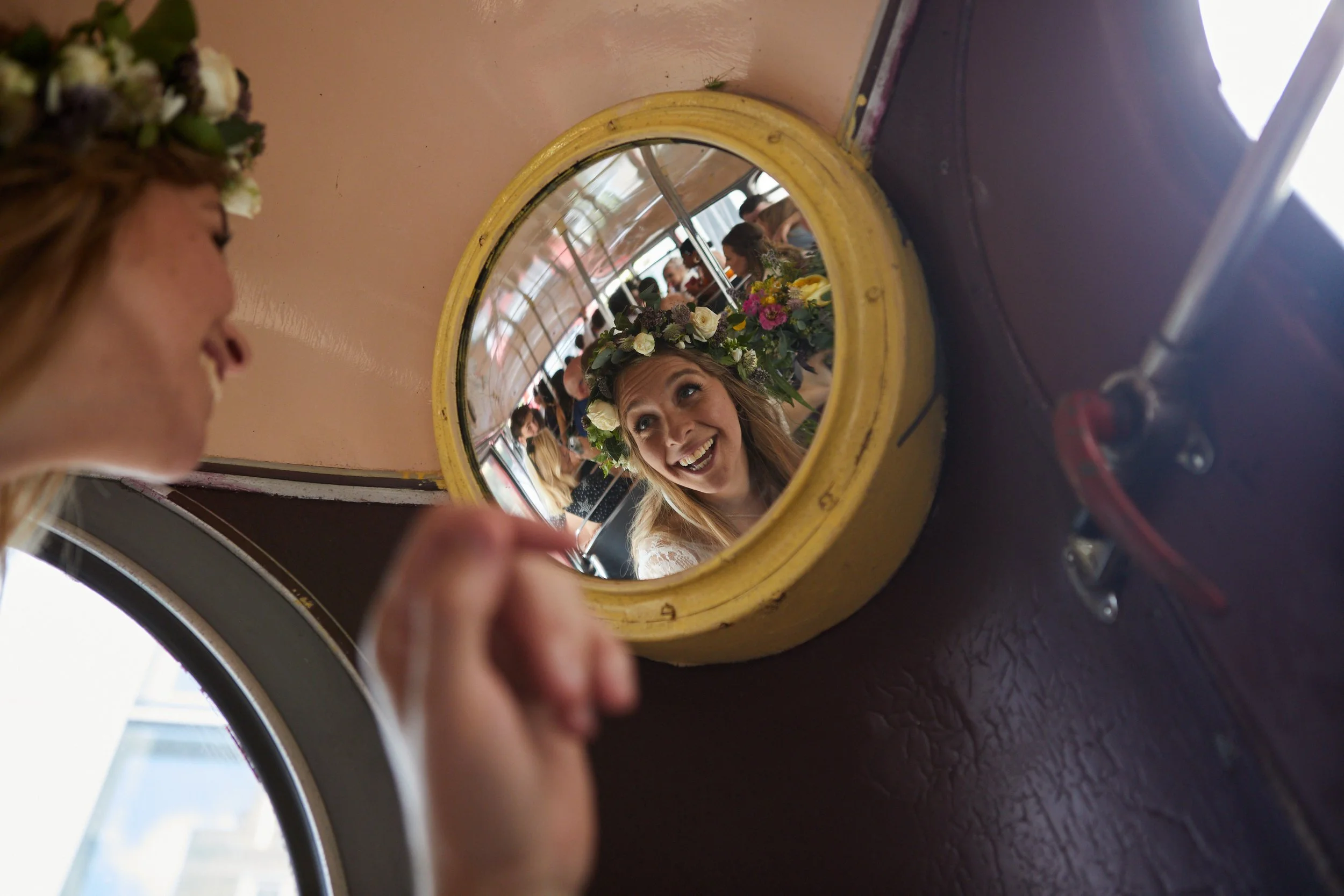 A woman with a floral crown is looking in a small round mirror, smiling happily, and the mirror reflects her face. The mirror is mounted on a wall inside a vintage vehicle, such as a bus or tram, with a rounded window visible in the background.