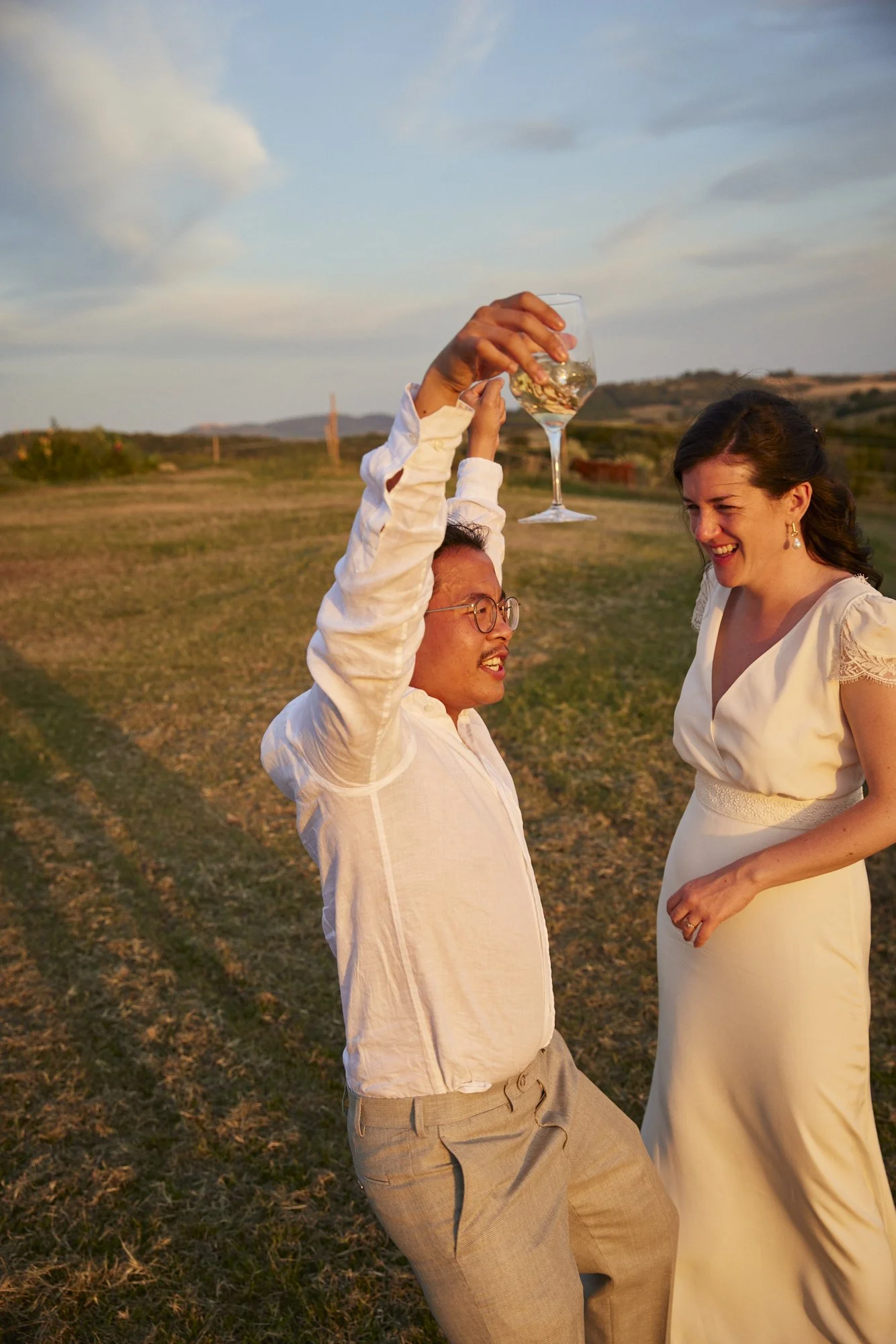 A man holding a glass of white wine up in celebration while a woman smiles and looks at him outdoors during sunset.