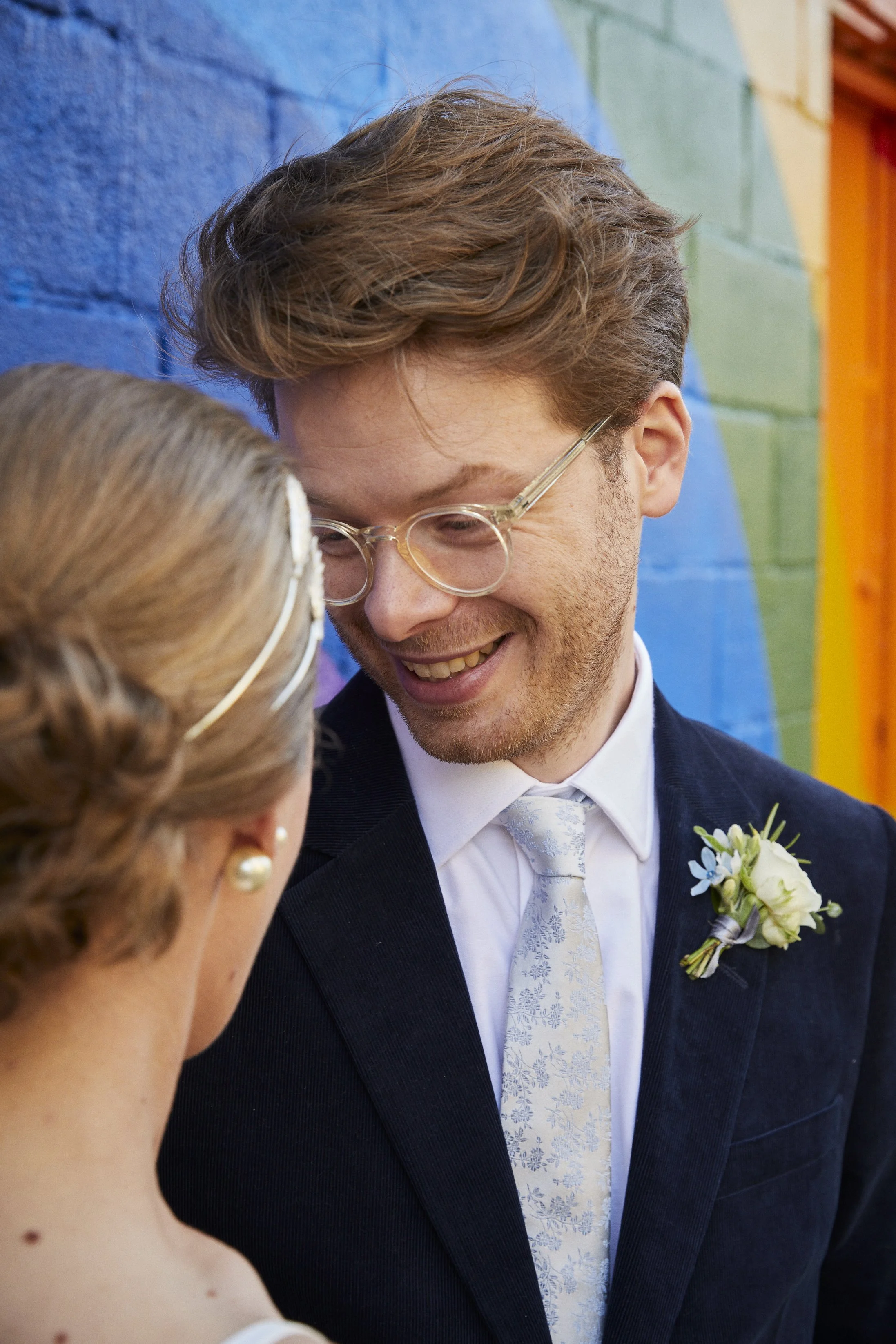 A man and woman in wedding attire smiling closely together against a colorful rainbow painted wall.