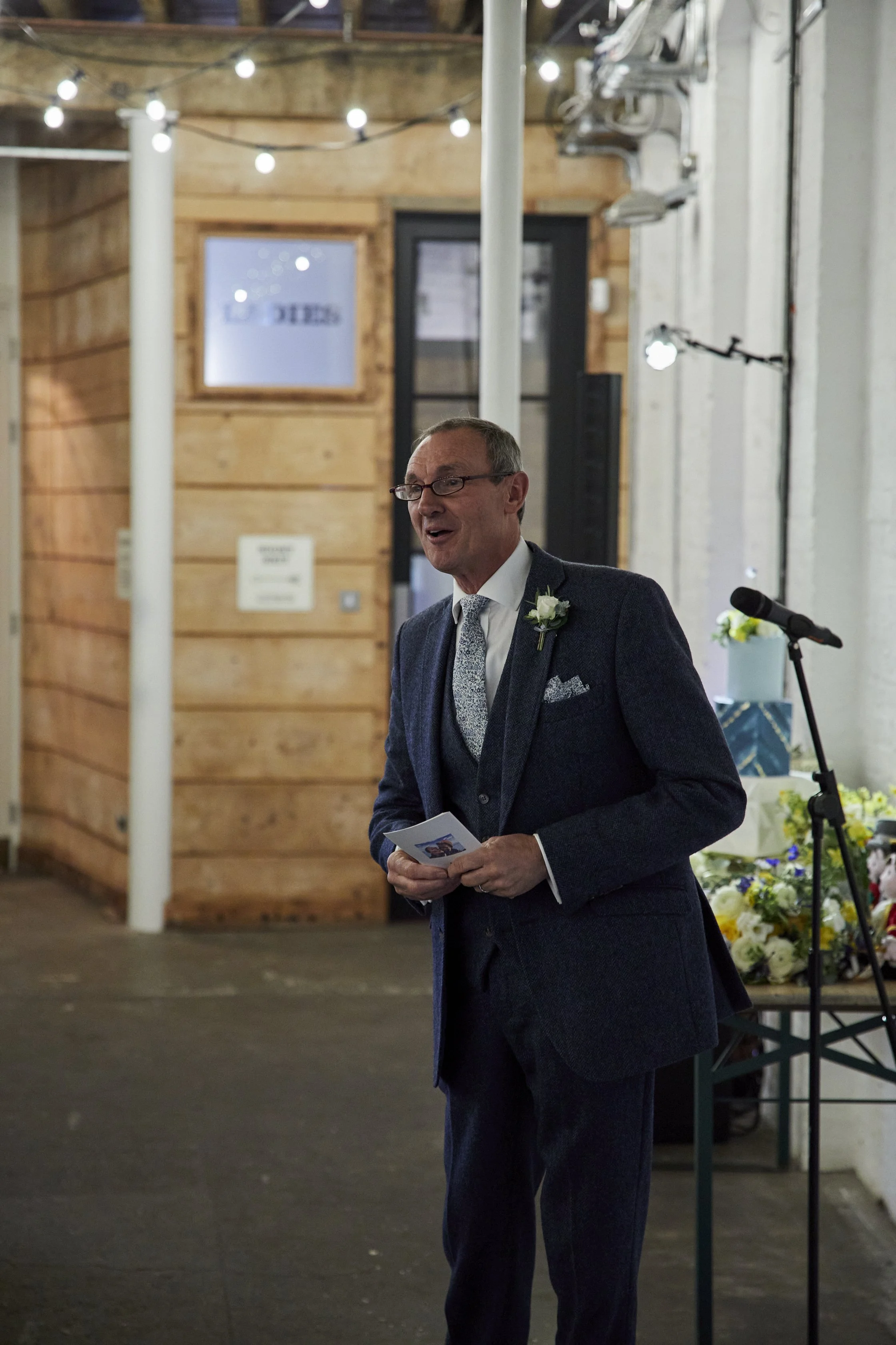 A man in a suit giving a speech at a wedding or formal event, with a microphone stand nearby, in a venue with wooden walls, string lights, and floral decorations.