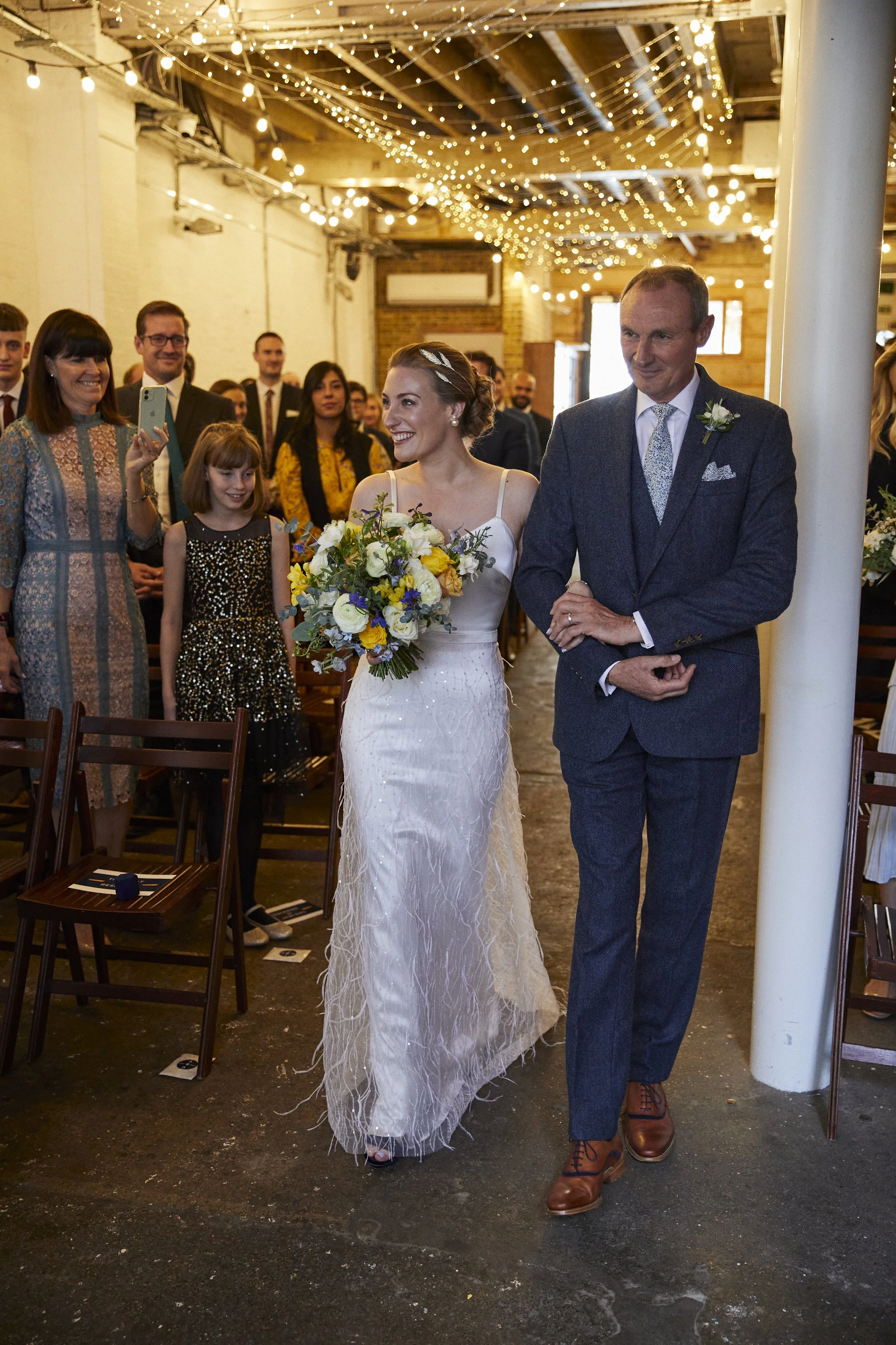Bride walking down the aisle with a man, likely her father, at a wedding ceremony. The bride holds a bouquet of flowers and is smiling. Guests stand on either side, some taking pictures, with string lights overhead in a rustic indoor venue.