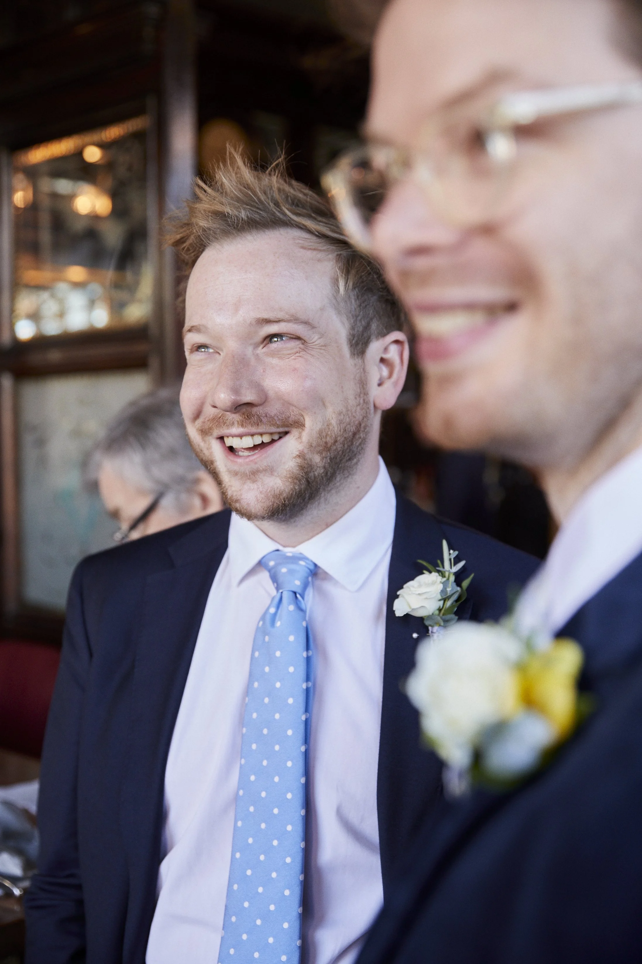 Two men in suits at a wedding, smiling and chatting, one wearing a light blue polka dot tie, both with white boutonnières