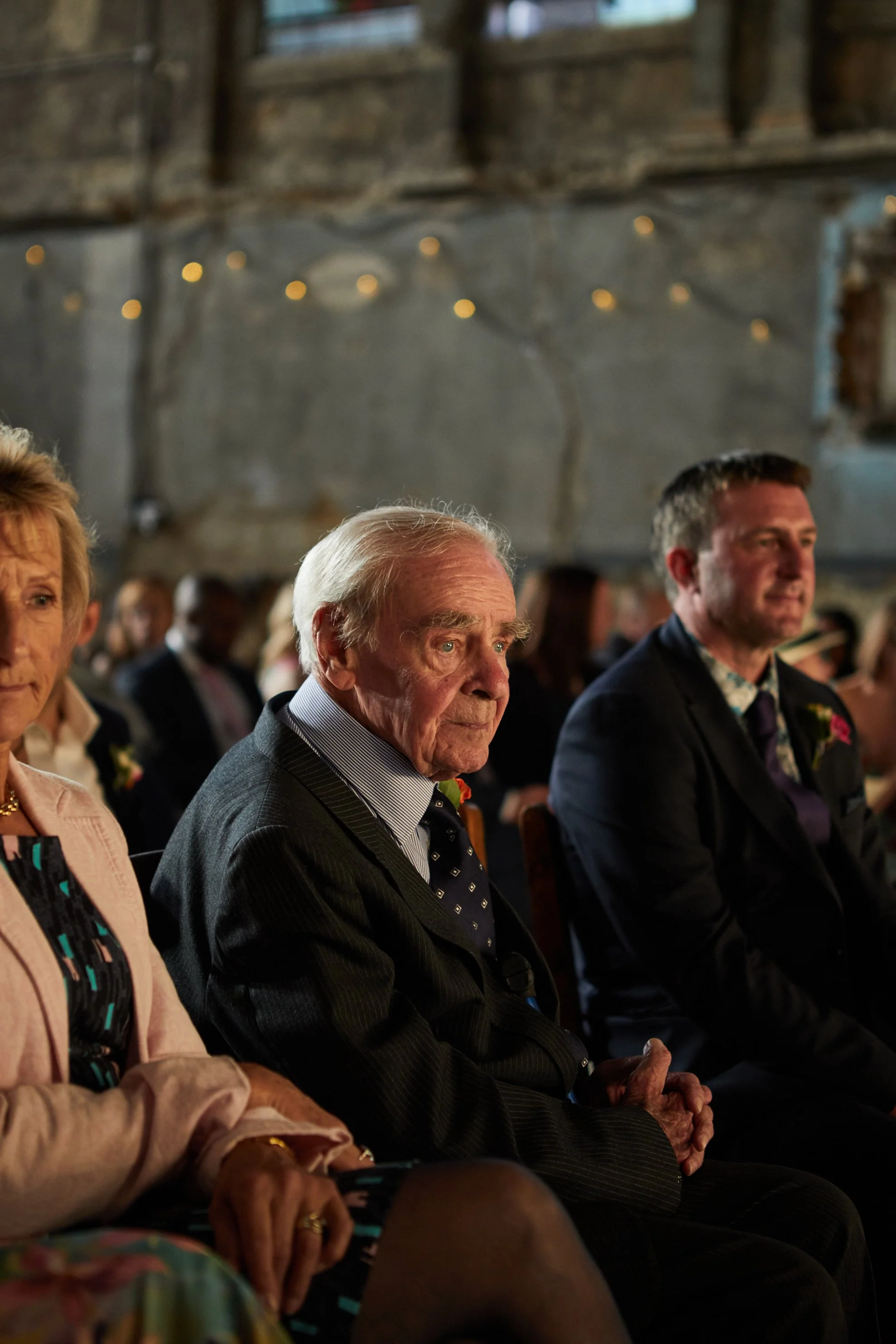 An elderly man and two other people seated at a formal event, possibly a wedding or ceremony, with dim lighting and string lights overhead.
