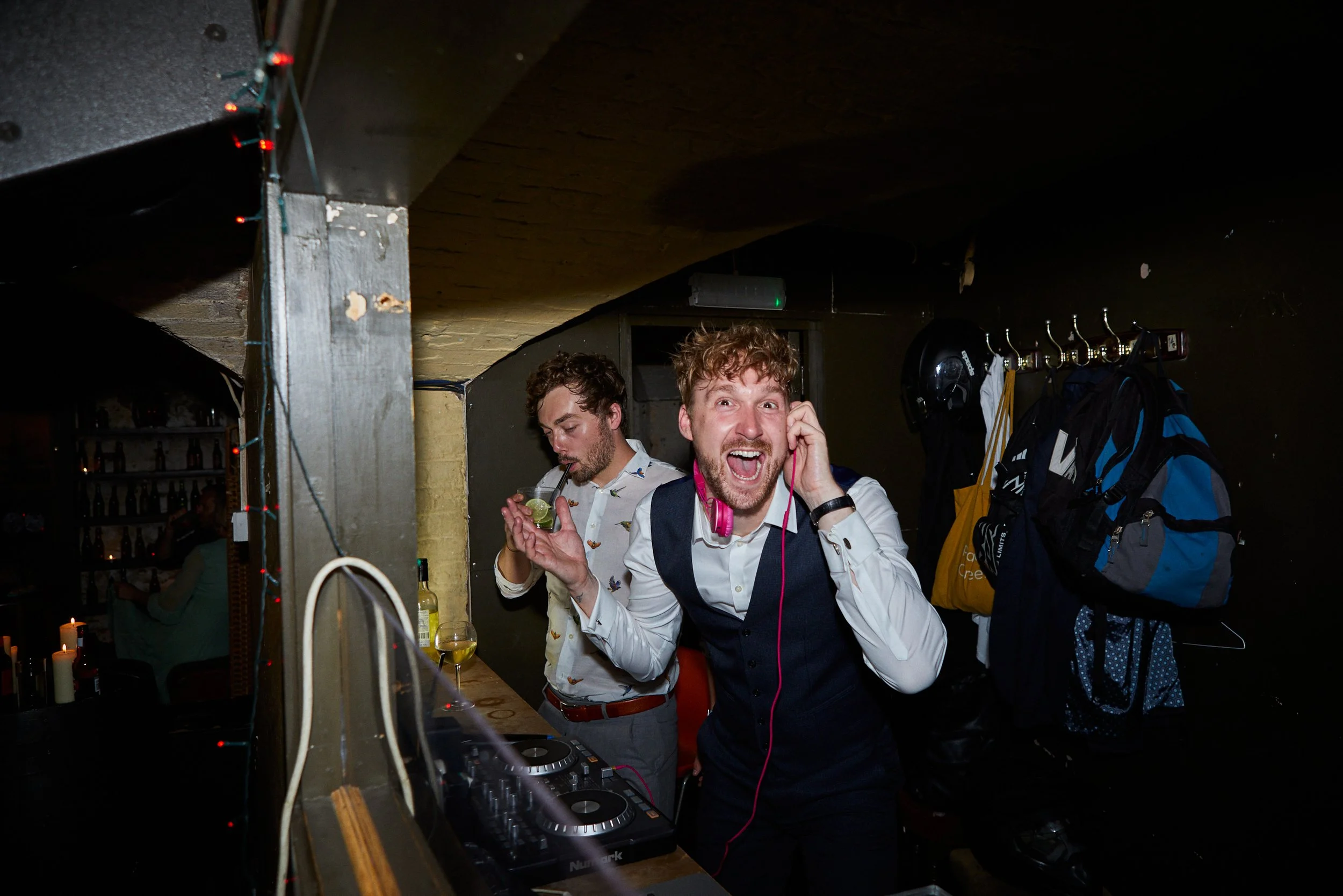 Two men at a party or bar, one DJ with pink headphones, smiling and making a cheerful pose, the other man drinking a green cocktail. The setting appears lively with bottles on shelves and party lights.