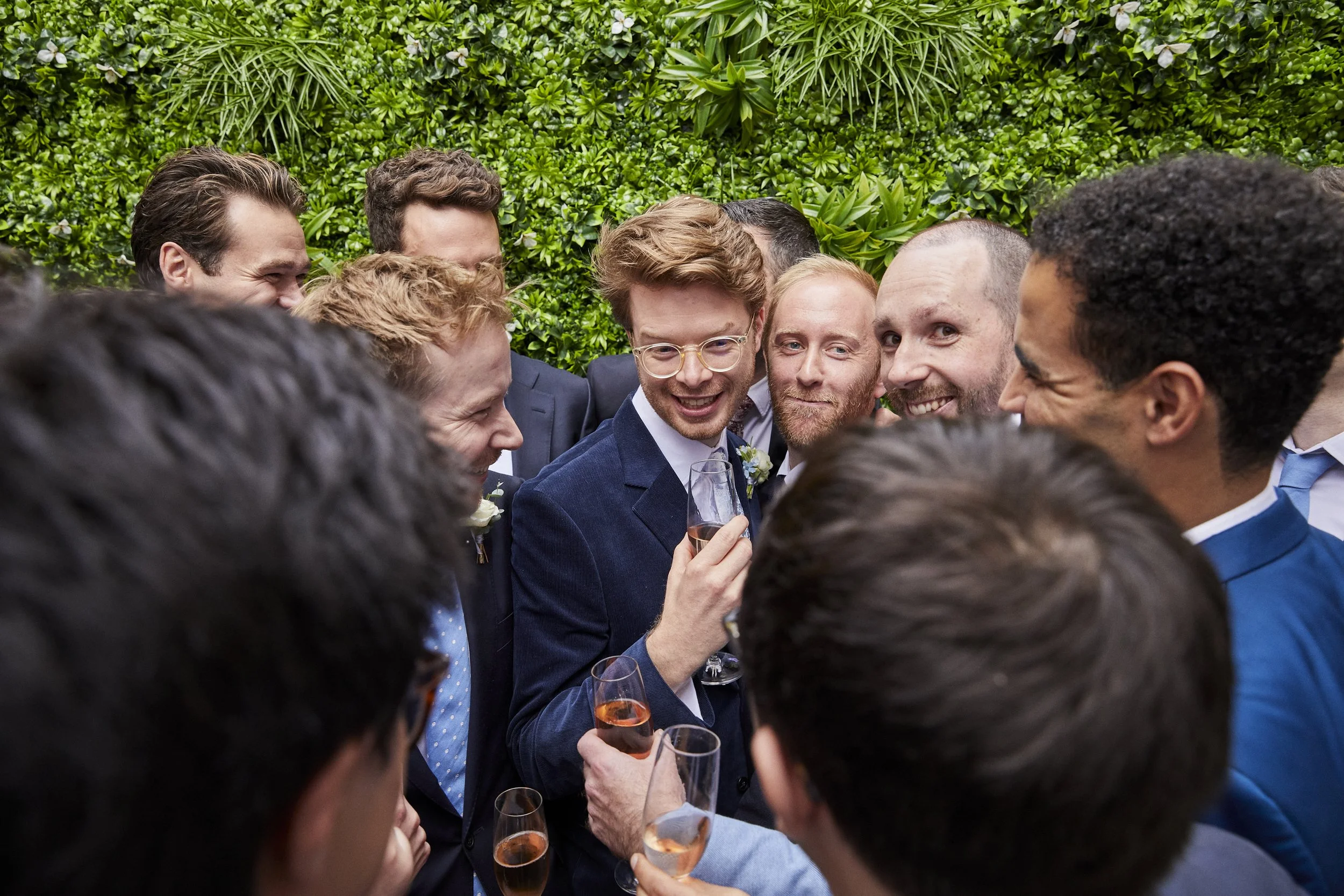 A group of men in suits and dresses in a semi-circle, smiling, with a person in the middle holding a glass, in front of a green leafy wall.