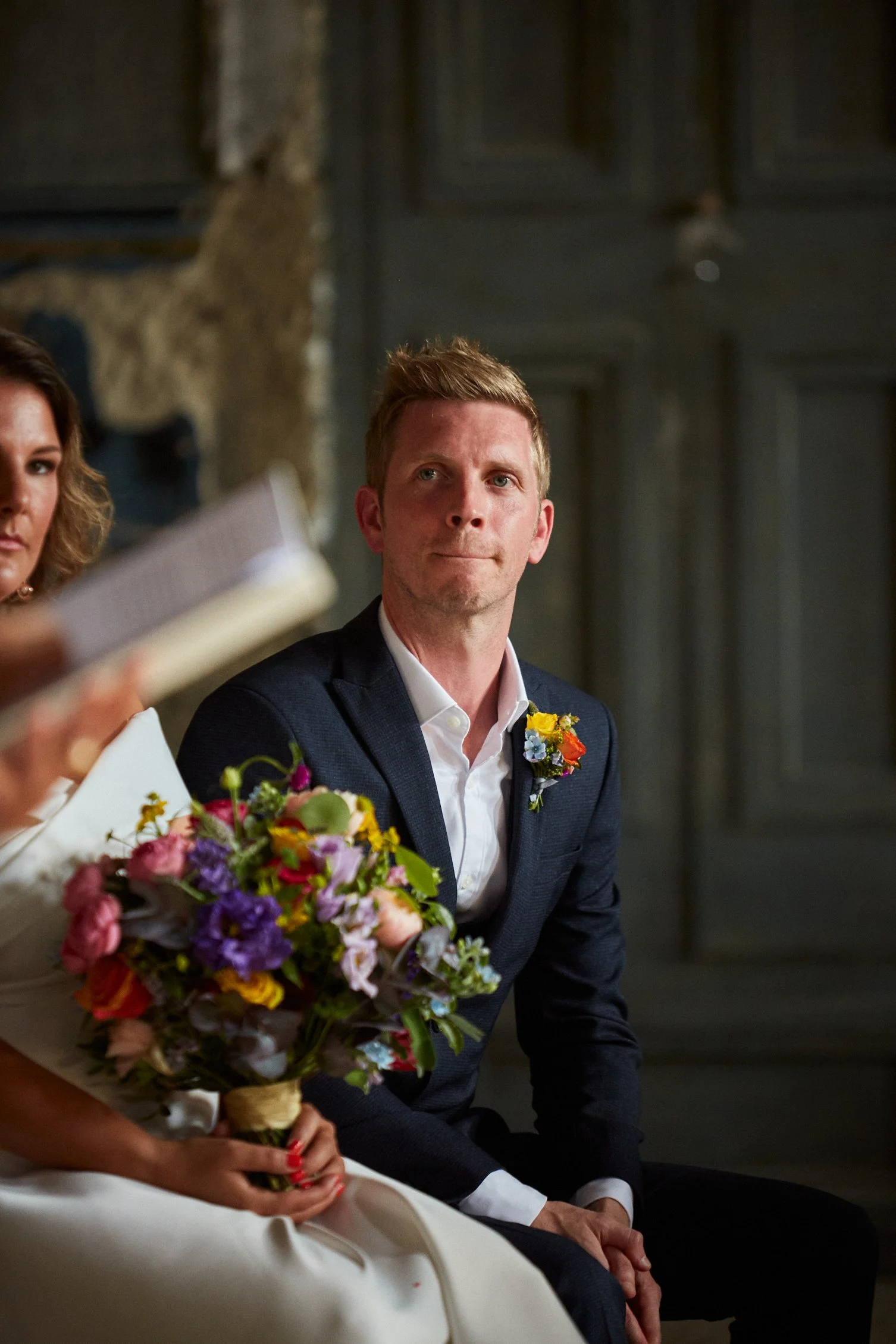 A man in a dark suit and white shirt sitting during a wedding ceremony, with a woman holding a colorful bouquet of flowers in front of him. The setting appears formal with a rustic background.