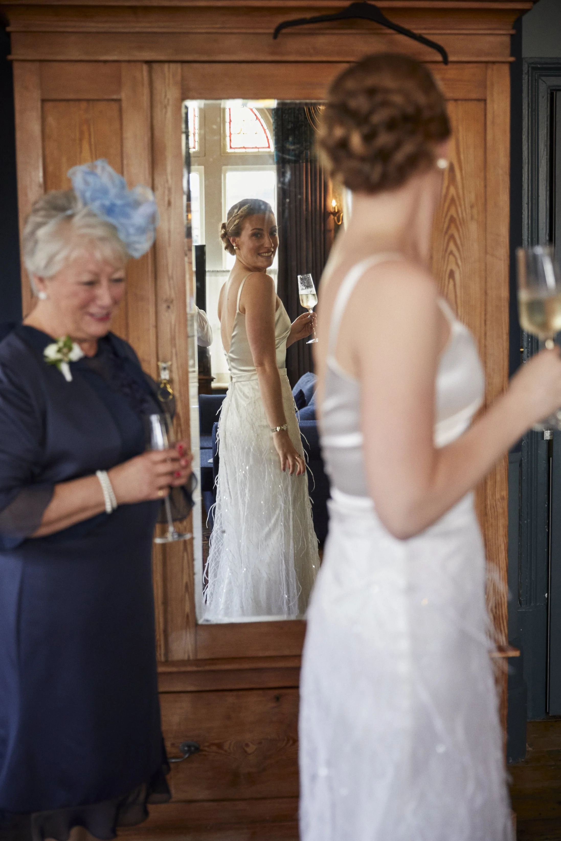 A bride in a white wedding dress looking at her reflection in a mirror, holding a glass of champagne, while an older woman in a dark dress and blue hat smiles and holds a glass of champagne nearby.