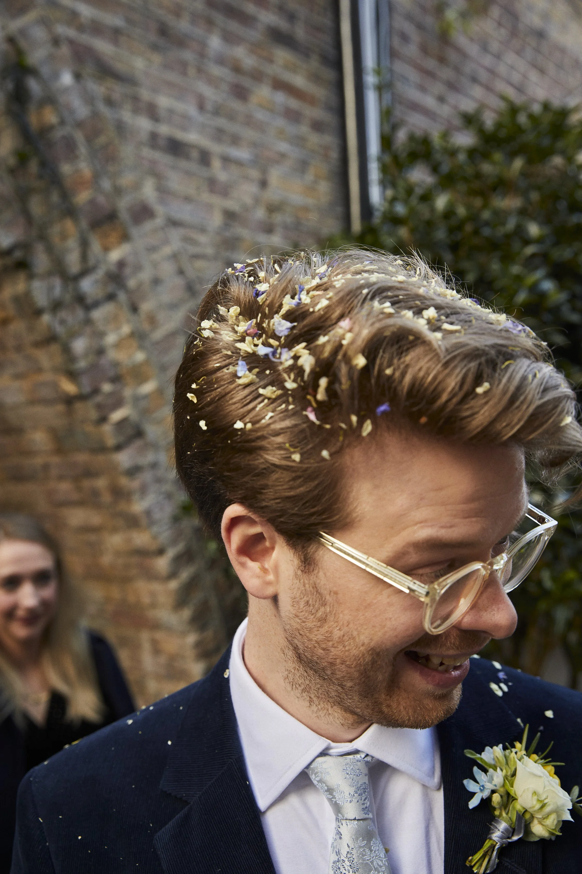 A man at a wedding with confetti in his hair, smiling and wearing glasses, a suit, tie, and boutonniere, outdoors by a brick wall.