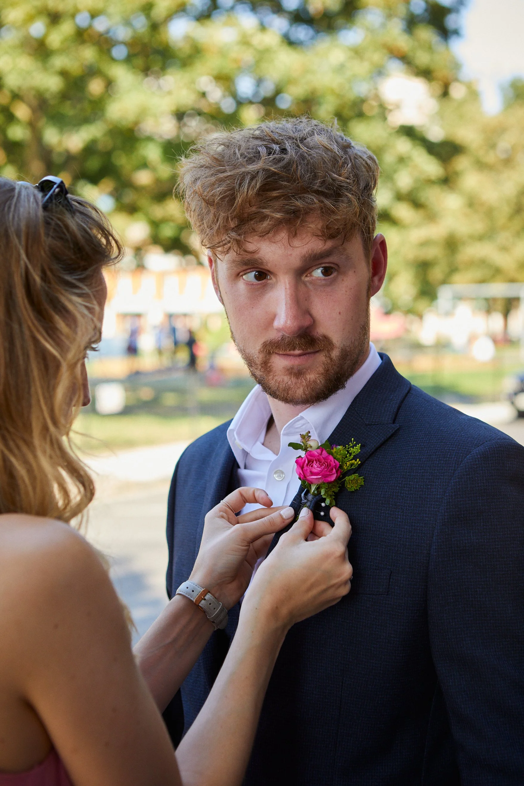 A woman pinning a pink boutonniere on a man's navy suit jacket during an outdoor event, with trees and people in the background.