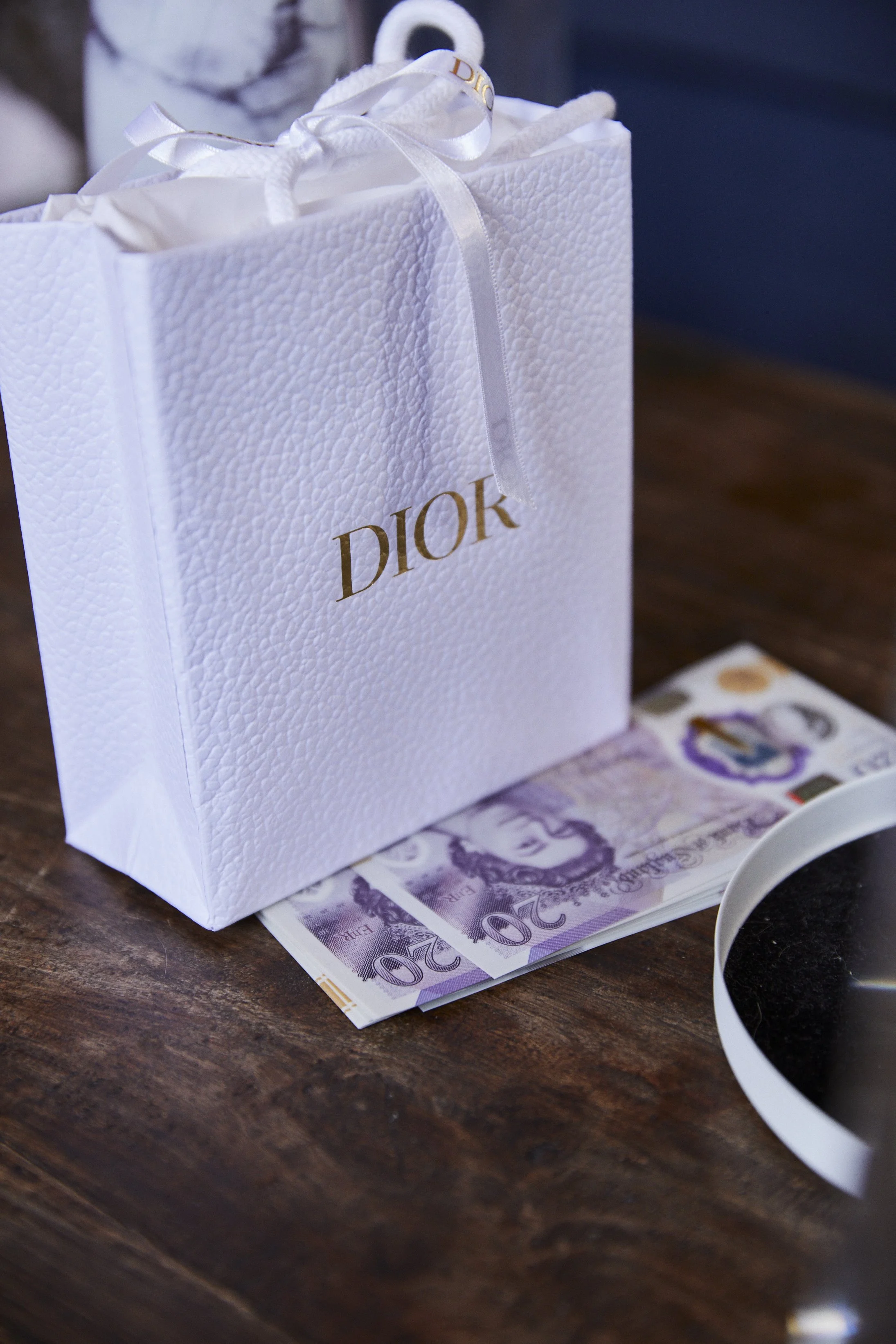 A white Dior gift bag with a ribbon handle sits on a dark wooden table, with some twenty pound banknotes partially underneath it.