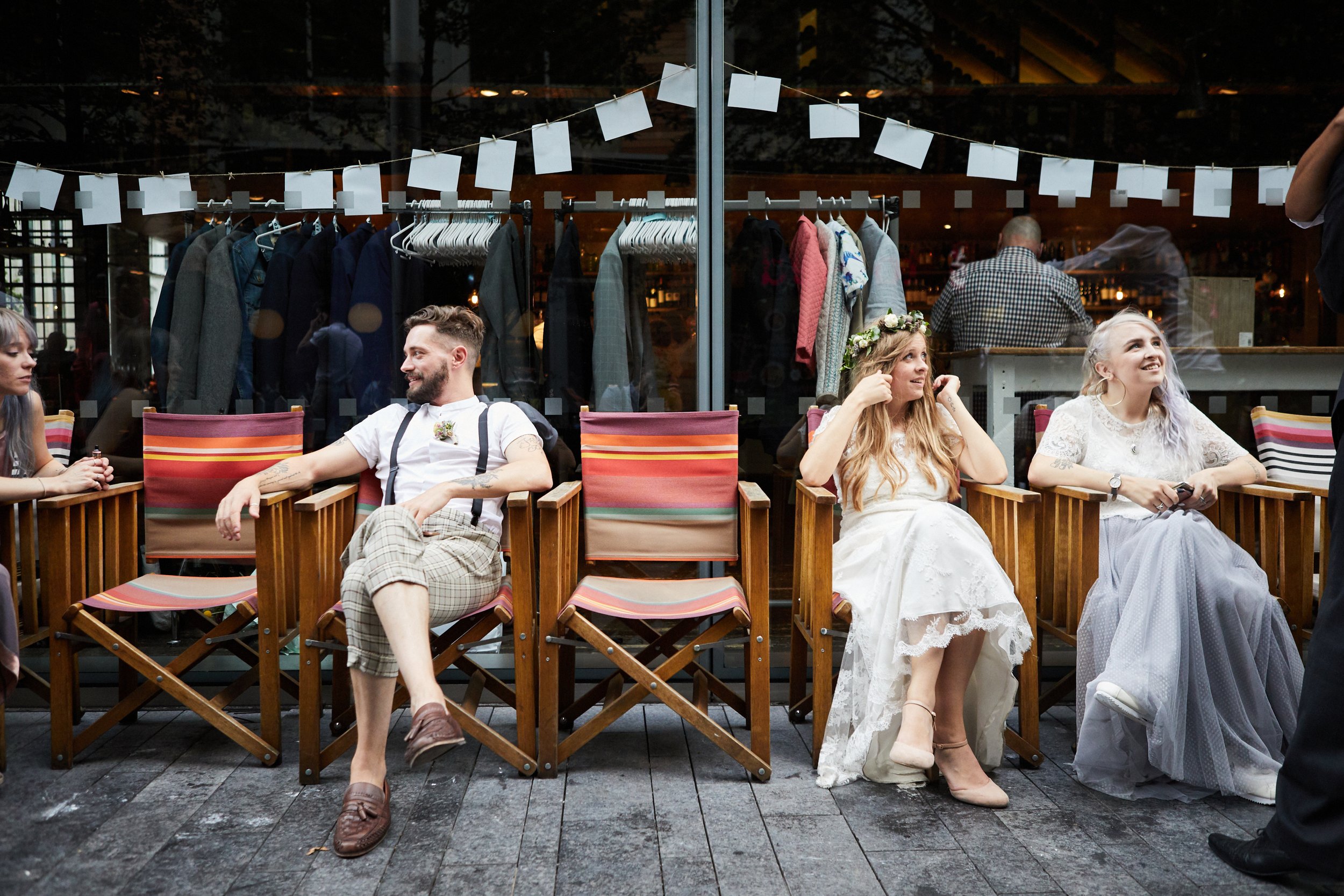 People sitting on colorful chairs outside a storefront with clothing hanging behind glass window.