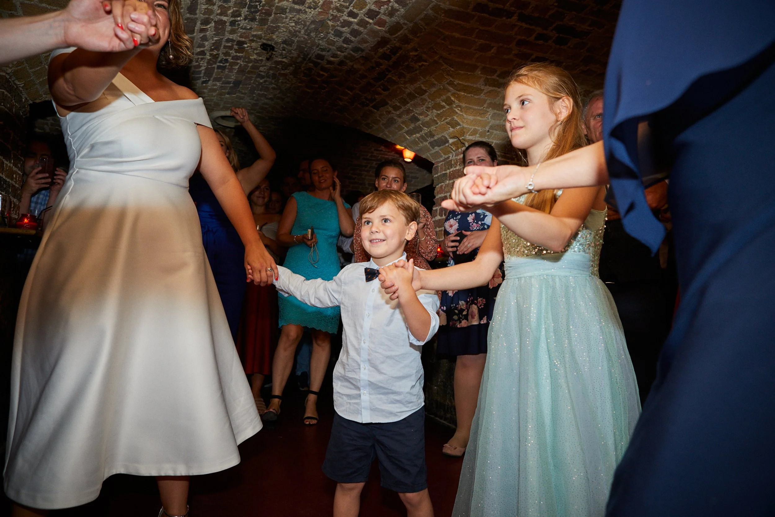 A young boy holding hands and dancing with women and girls in a celebration or wedding reception in a brick-walled venue.