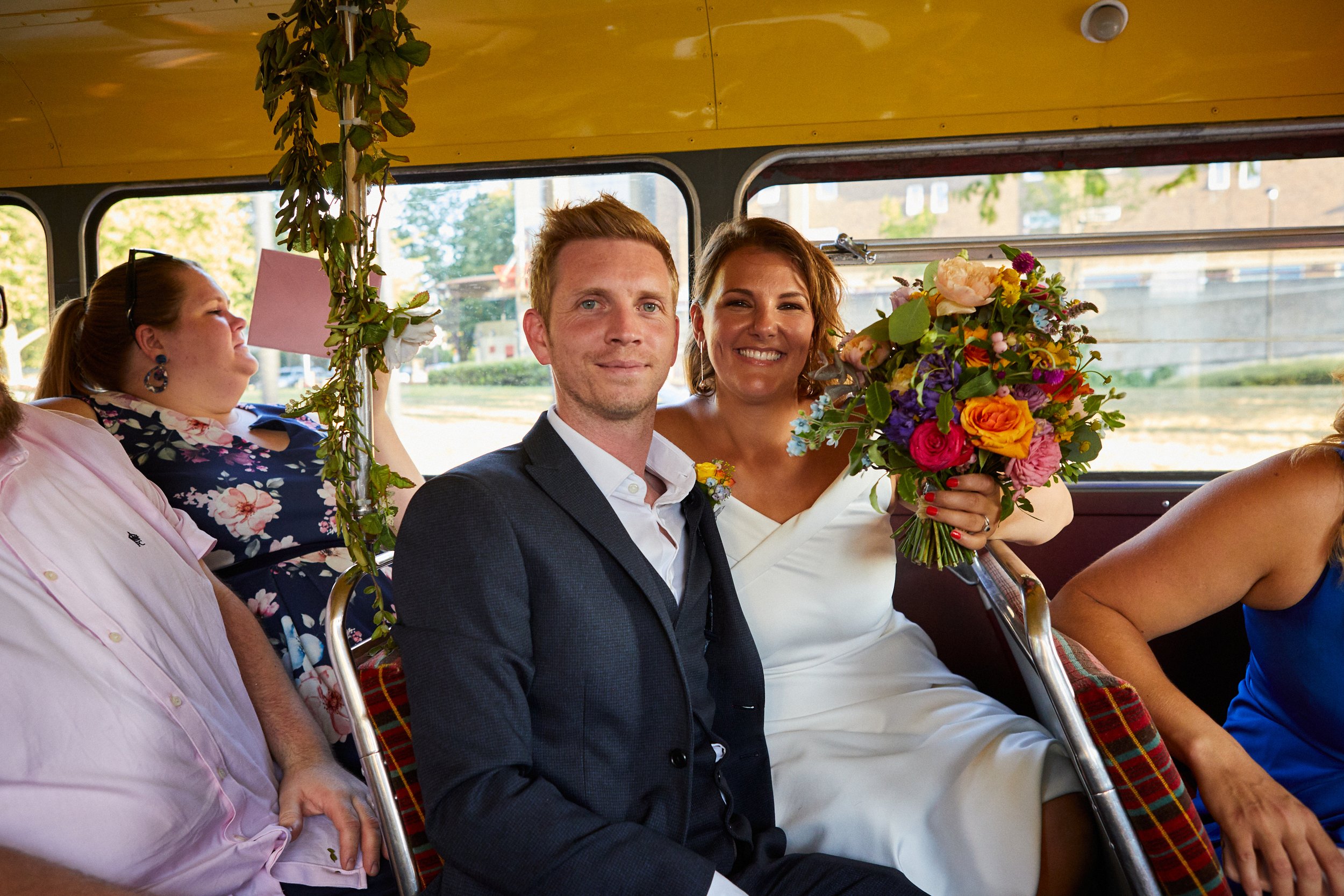 A smiling couple on a bus, with the woman holding a large colorful bouquet of flowers. The man is dressed in a dark suit, and the woman is in a white dress. There are other people seated around them, and a window showing a street scene outside.