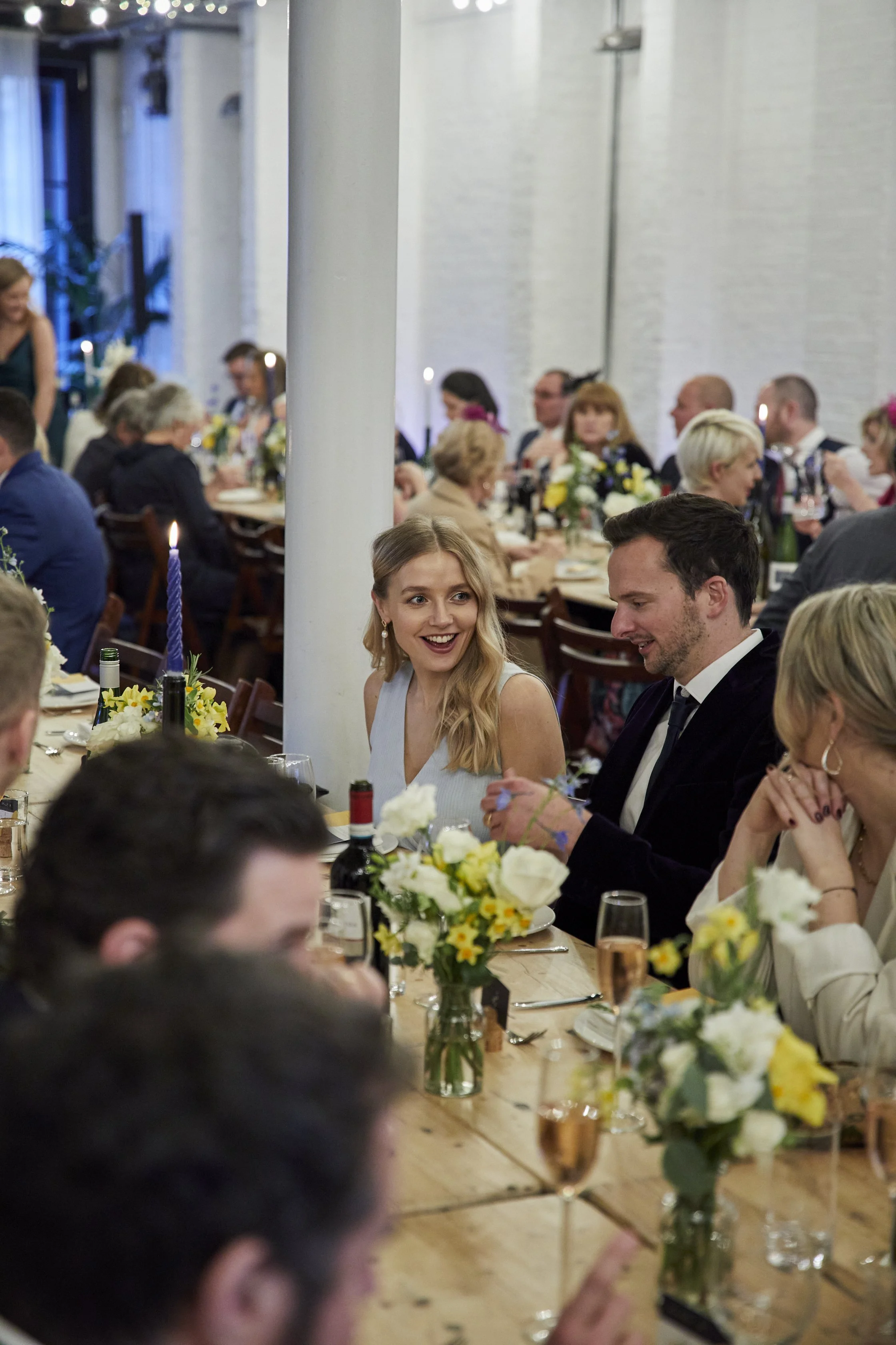 People celebrating at a wedding reception, sitting at a long table decorated with flowers, bottles of wine, and candles.
