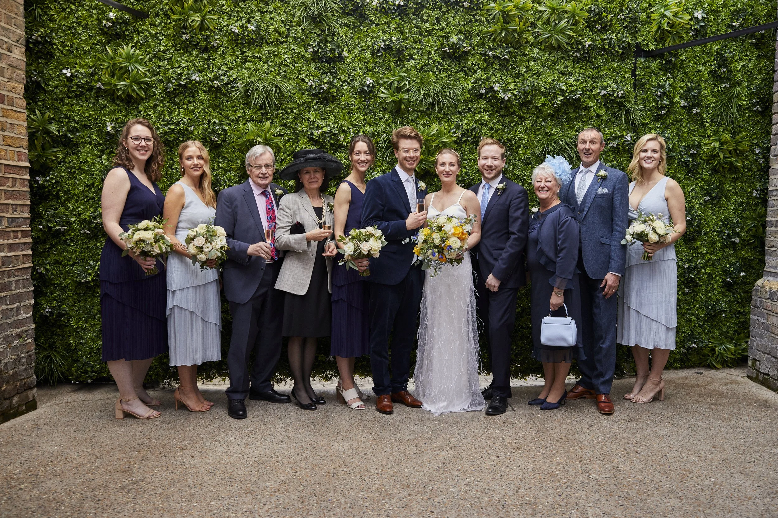 A group of people at a wedding posing in front of a lush green wall, with women holding bouquets and men in suits.