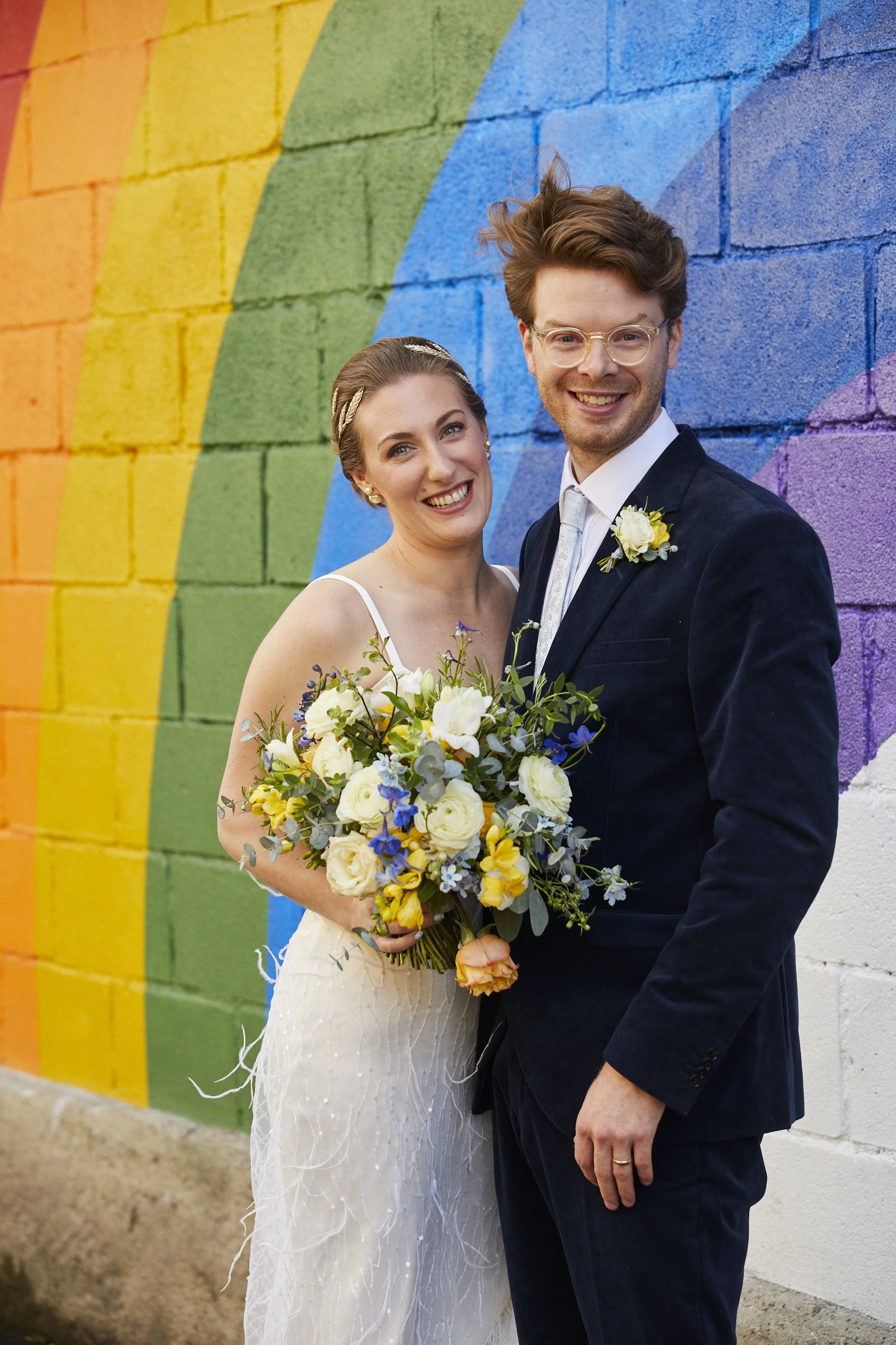 A smiling bride and groom standing in front of a colorful rainbow mural wall, holding a bouquet of white, yellow, and purple flowers. The bride is wearing a white dress with spaghetti straps, and the groom is dressed in a dark suit with a white shirt