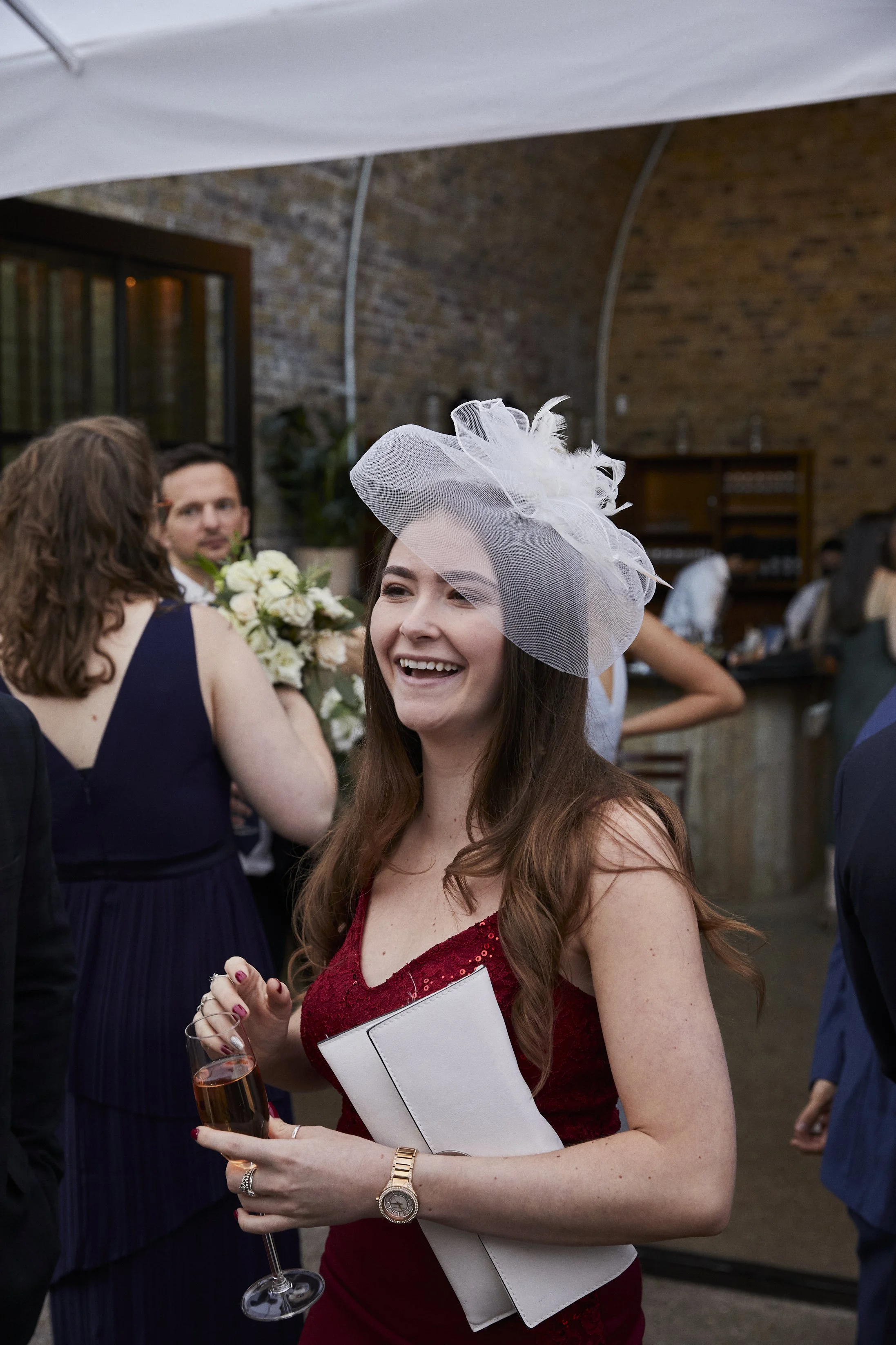 A smiling woman in a red dress wearing a large white fascinator hat, holding a glass of champagne and a white clutch, at a social event or party.