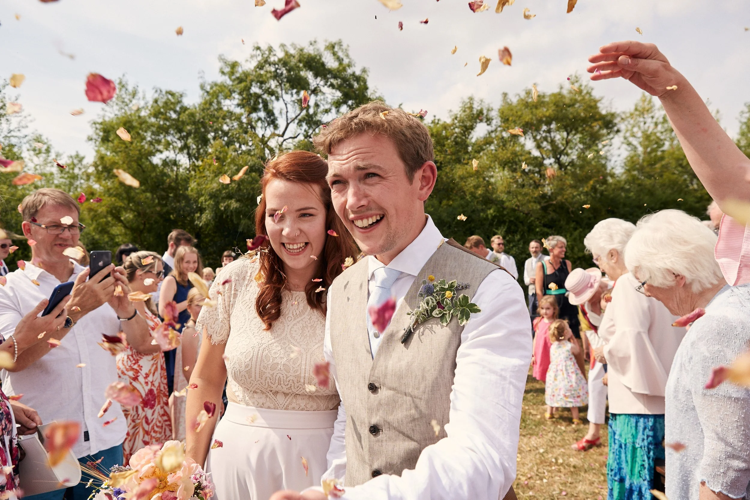 A joyful wedding celebration outdoors with guests throwing flower petals at a smiling bride and groom, surrounded by trees and a blue sky.