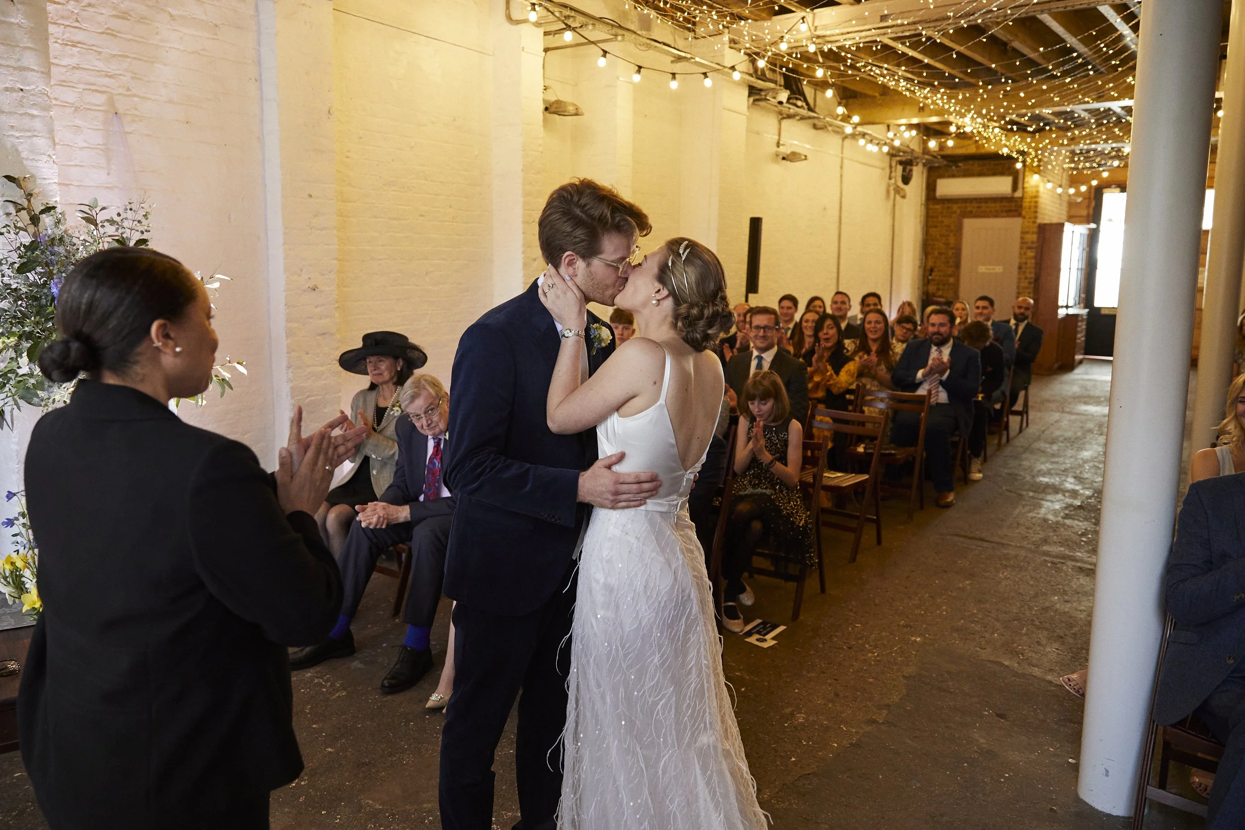 A bride and groom share a kiss during their wedding ceremony inside a decorated venue with string lights on the ceiling, with guests clapping and watching in the background.