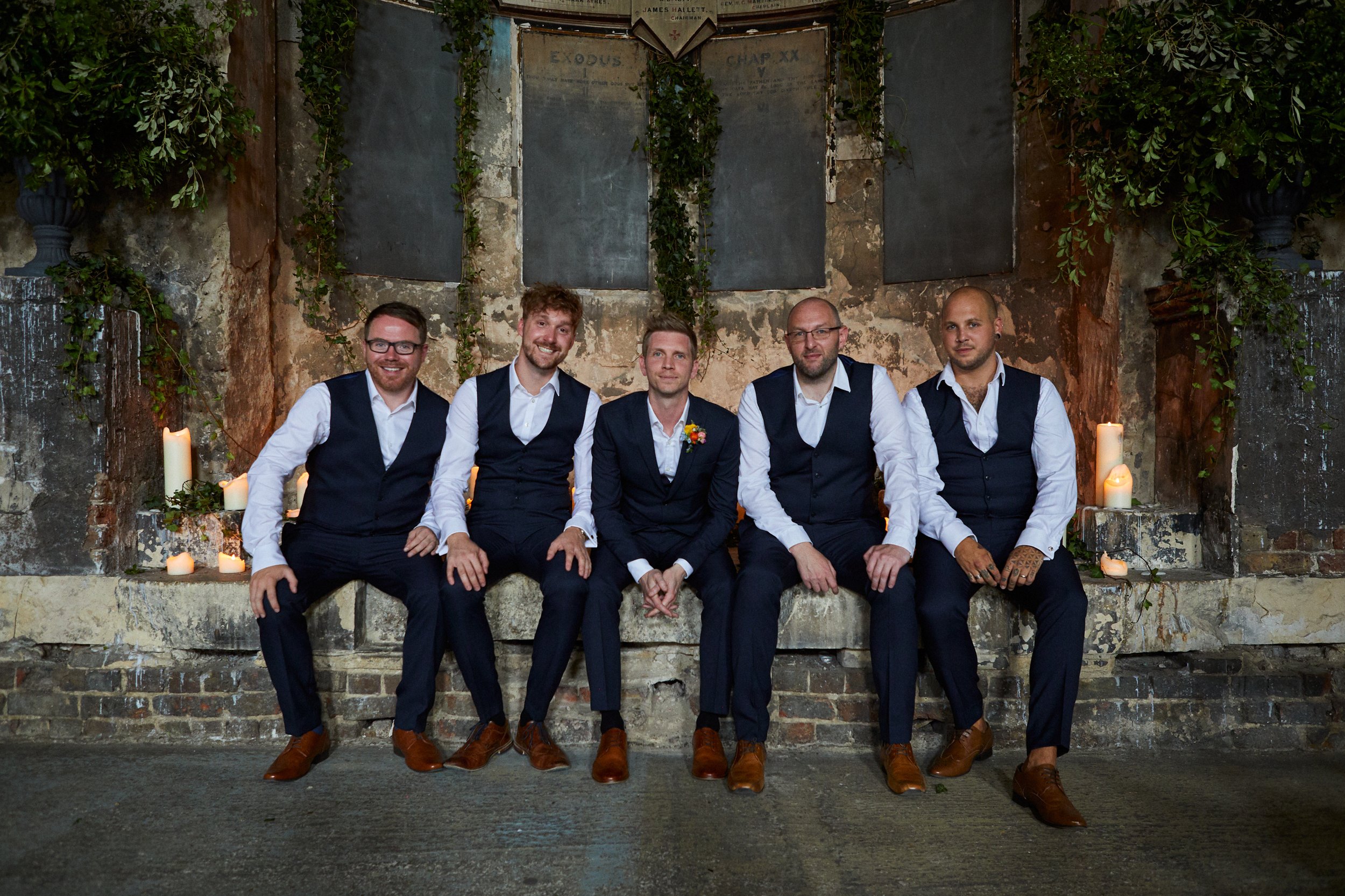 A group of six men dressed in formal attire, sitting on a stone bench in front of a rustic wall with candles and greenery, during a wedding or formal event.