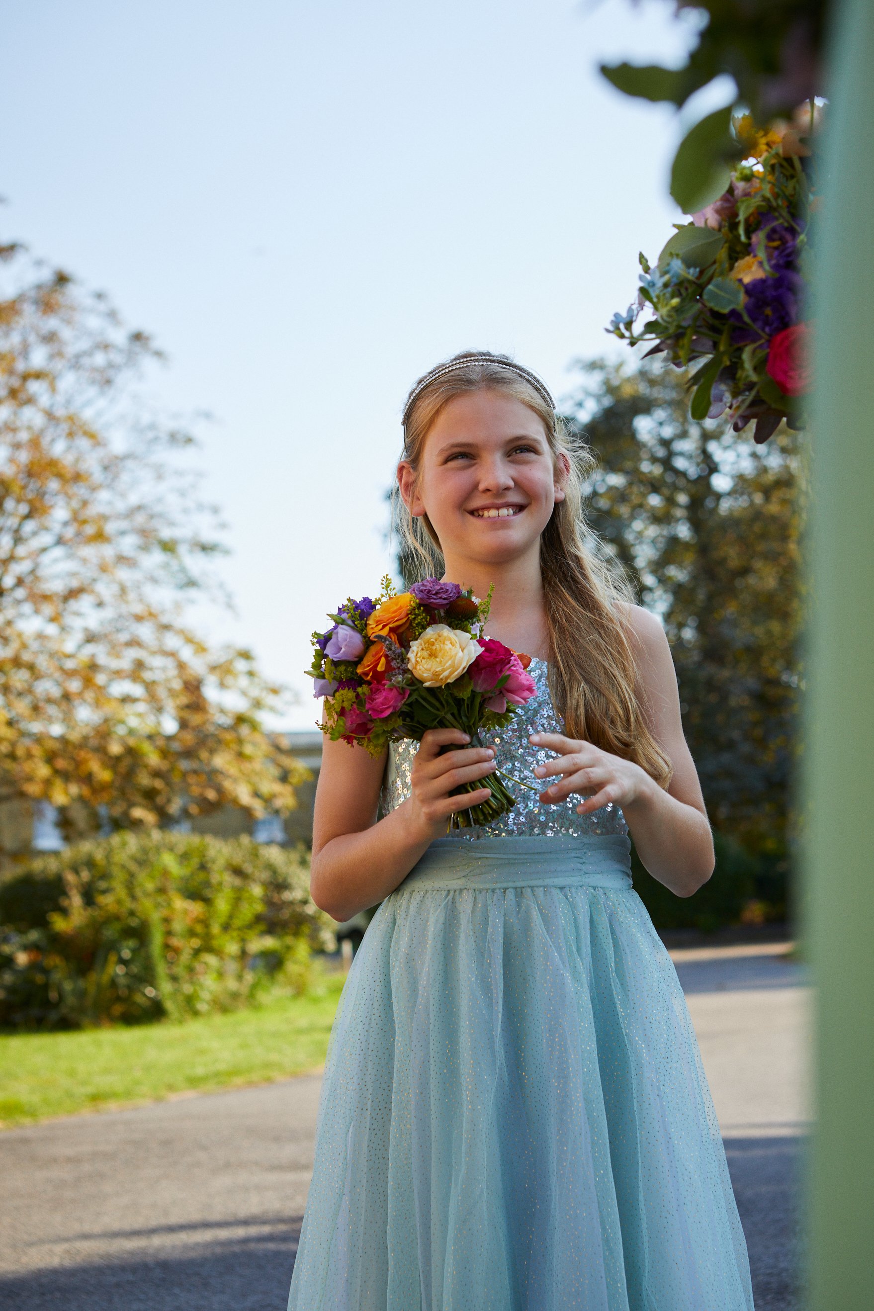 A young girl in a sparkly light blue dress smiling while holding a colorful bouquet of flowers outdoors on a sunny day.