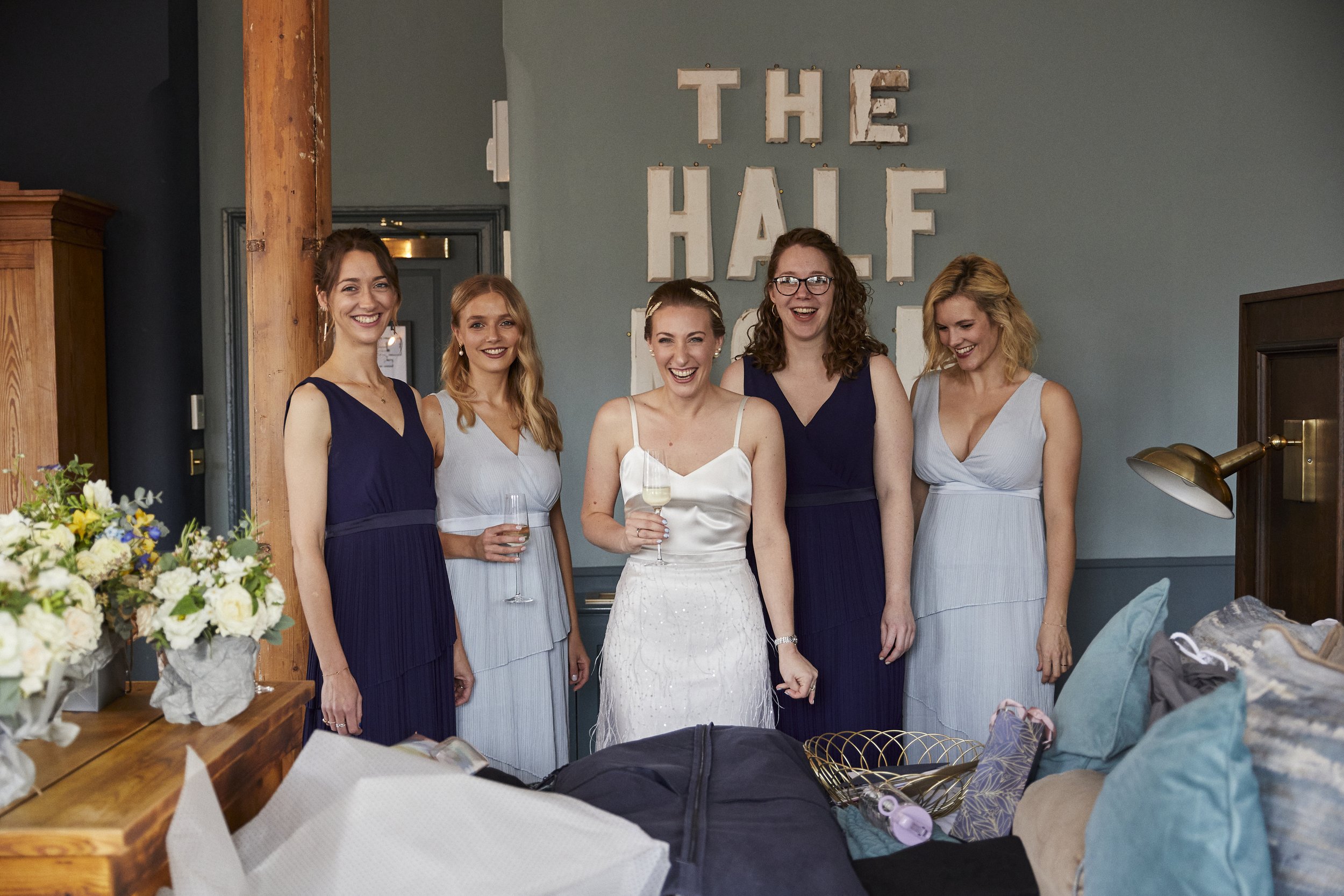 Group of six women in dresses smiling and celebrating, with the words 'The Half' on the wall behind them.