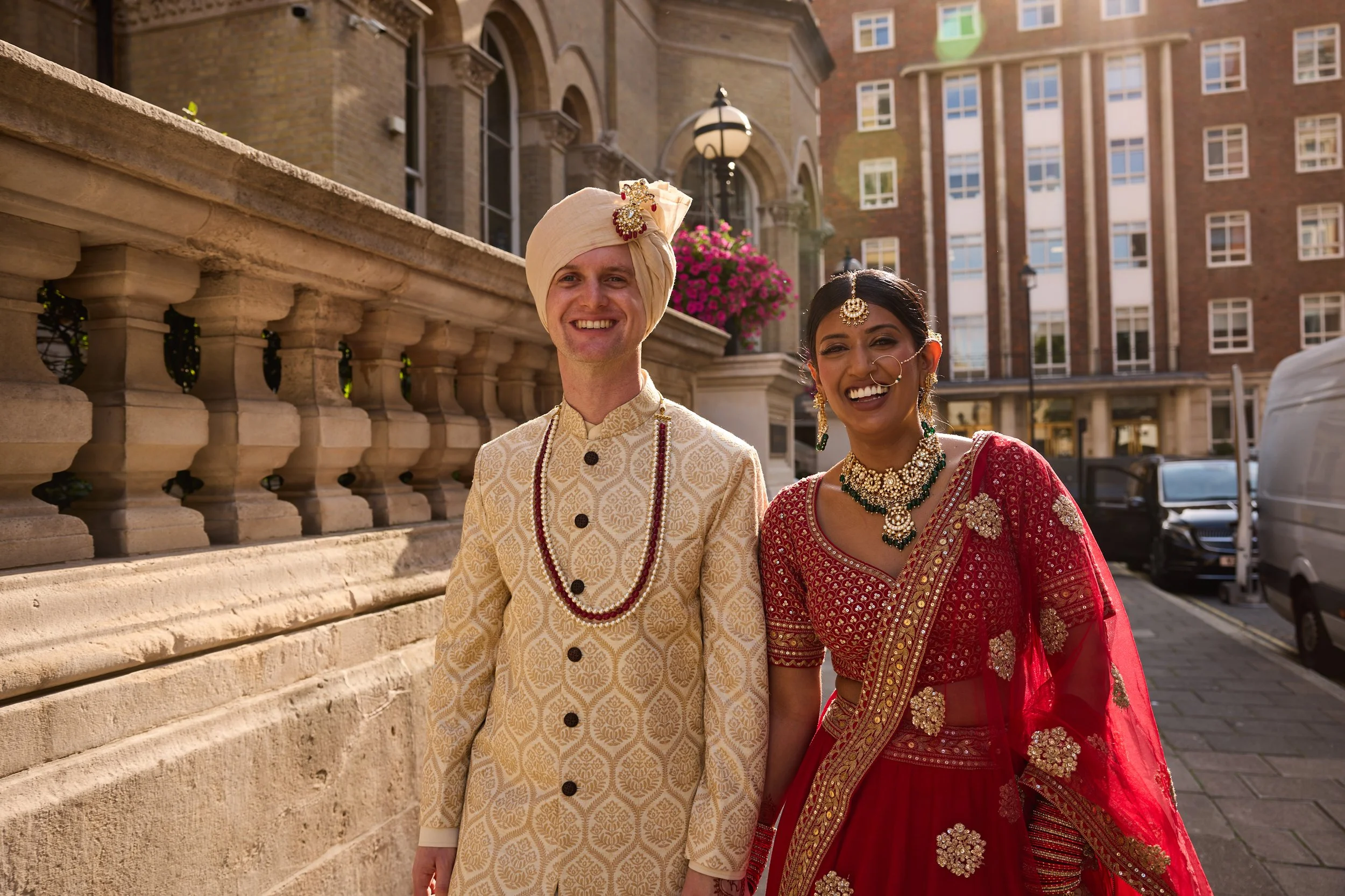 A smiling couple dressed in traditional Indian wedding attire standing outdoors in an urban setting with brick buildings and parked cars in the background.
