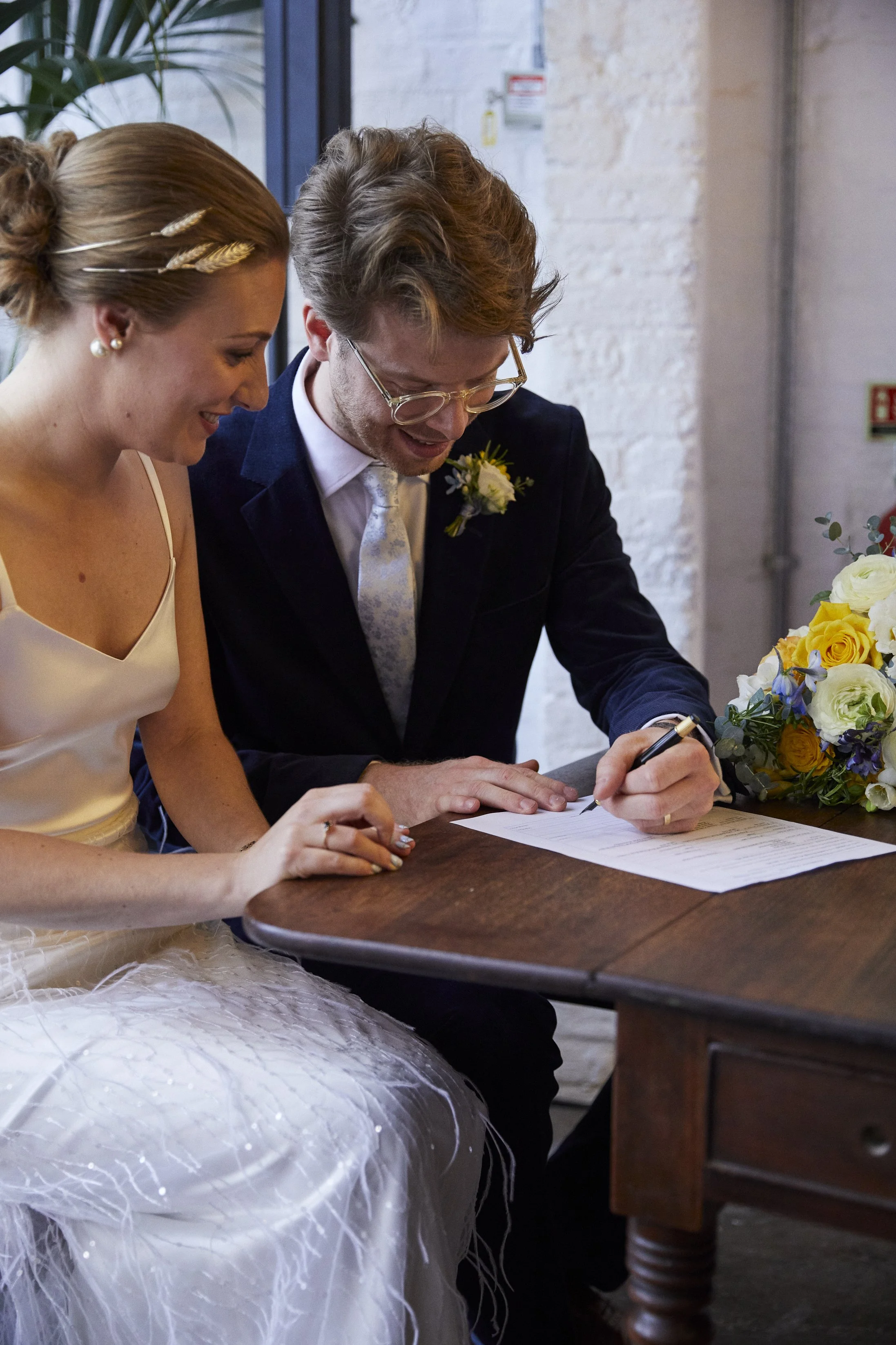 A bride and groom signing their marriage certificate at a wedding celebration, with a bouquet of flowers nearby.