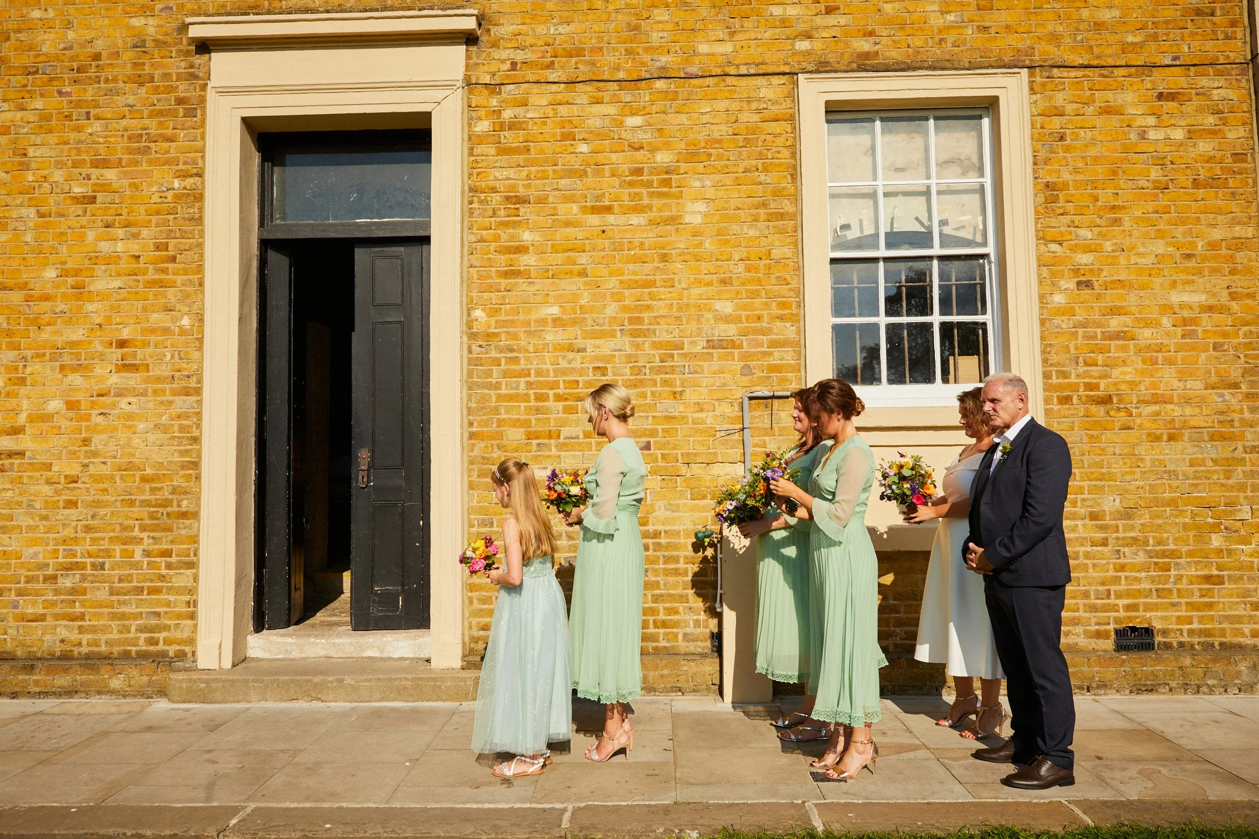 People lined up outside a building with a yellow brick wall, waiting for an event, possibly a wedding, holding bouquets of flowers.