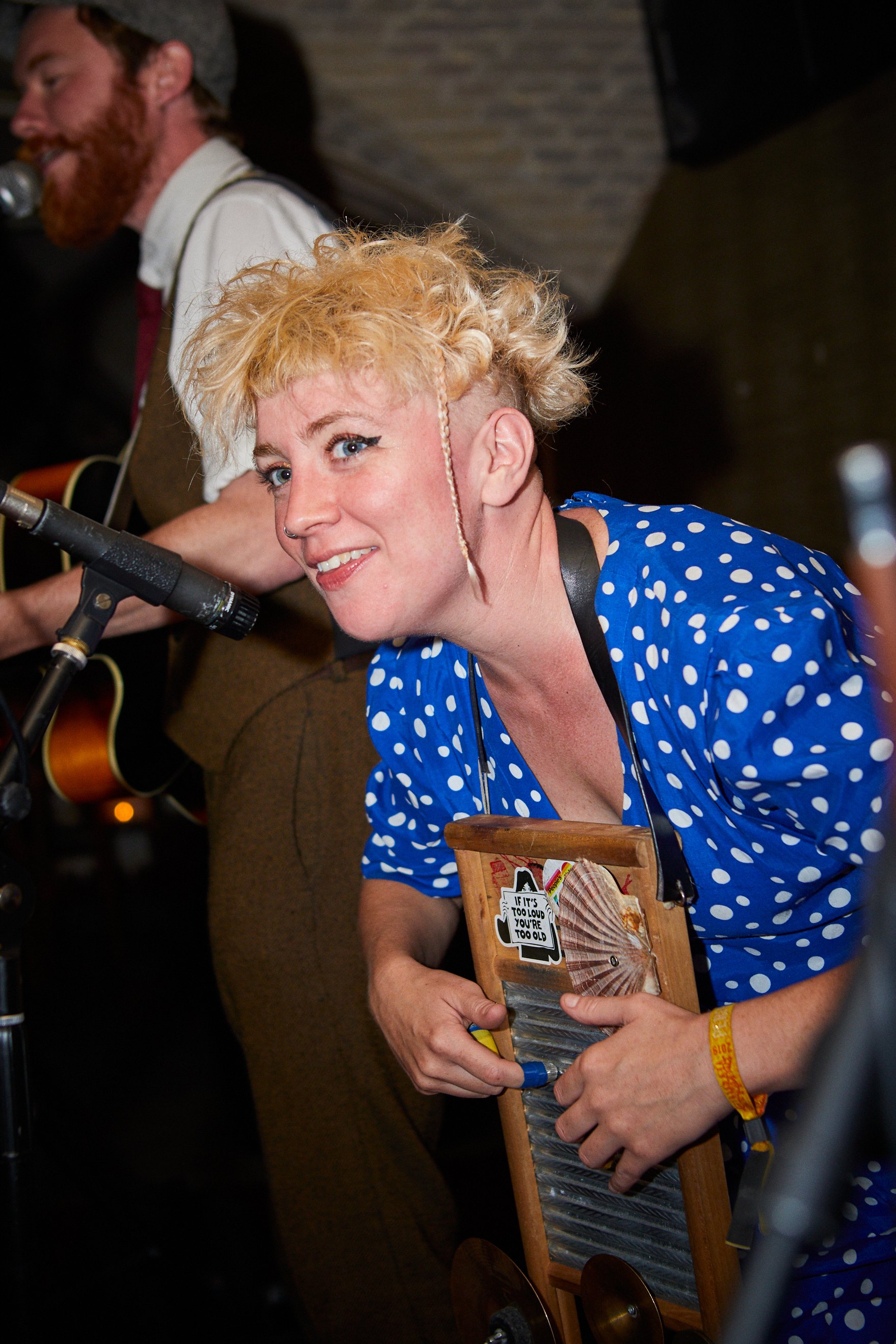 A woman with short, curly blonde hair, blue eyes, and freckles, wearing a blue polka-dot shirt, smiling and posing with a washboard. Behind her, a man with a beard and mustache, playing an acoustic guitar, is partly visible. They are performing in a 