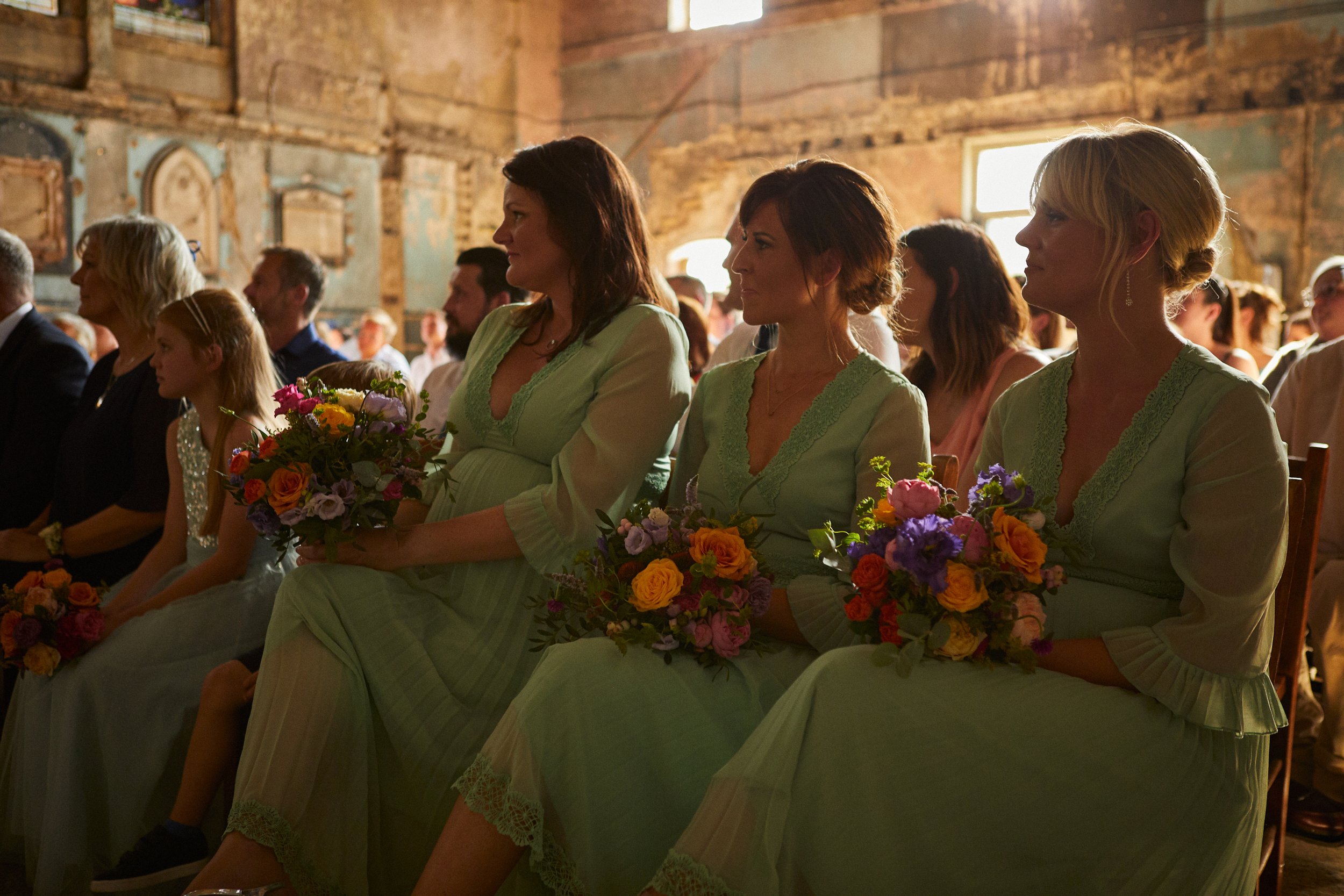 Women in light green dresses sitting in a rustic, warmly lit venue, holding colorful floral bouquets during a wedding or special event.