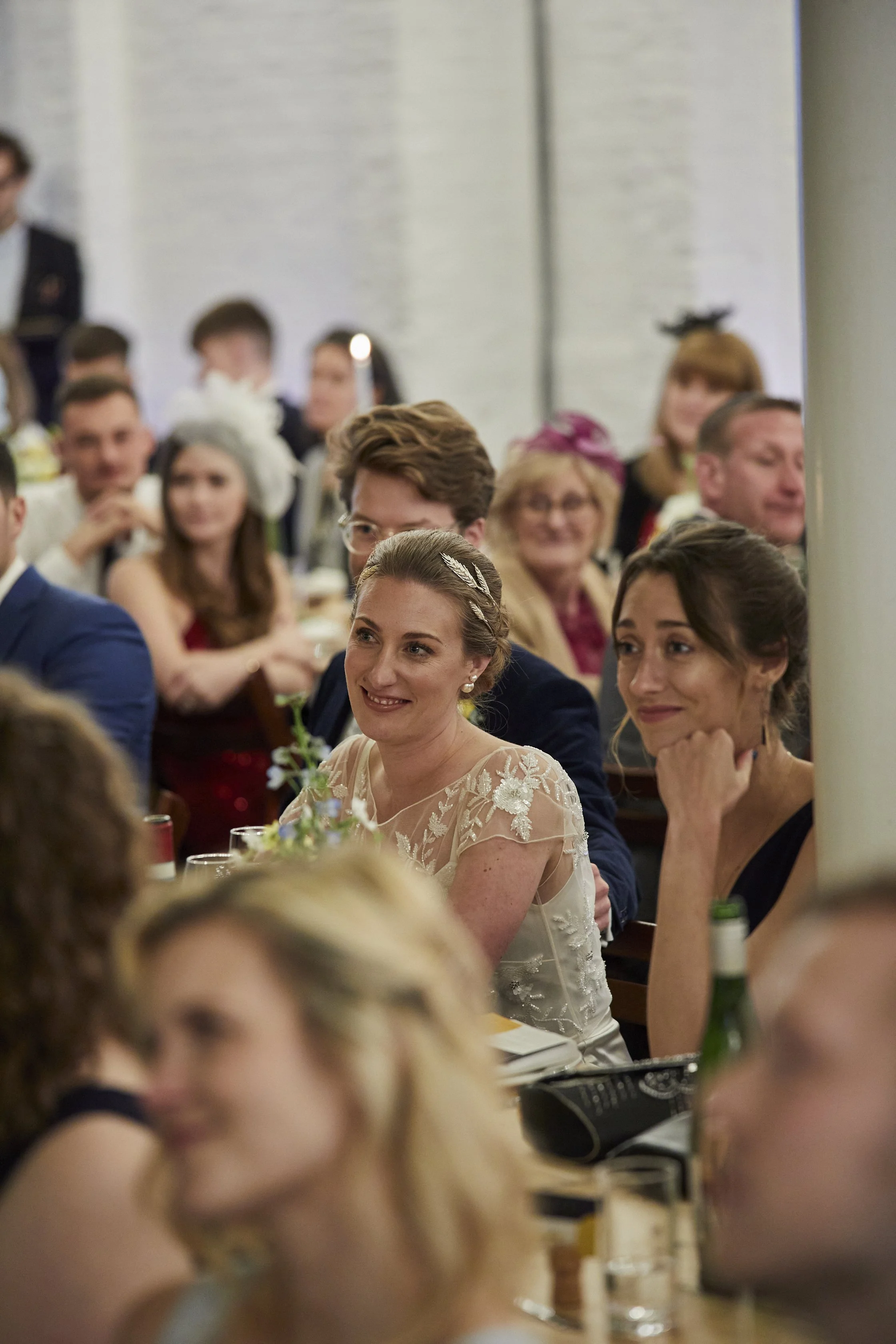 Guests seated at tables during a wedding reception, including a woman in a lace wedding dress and a woman in a black dress, with a diverse group of people in the background.