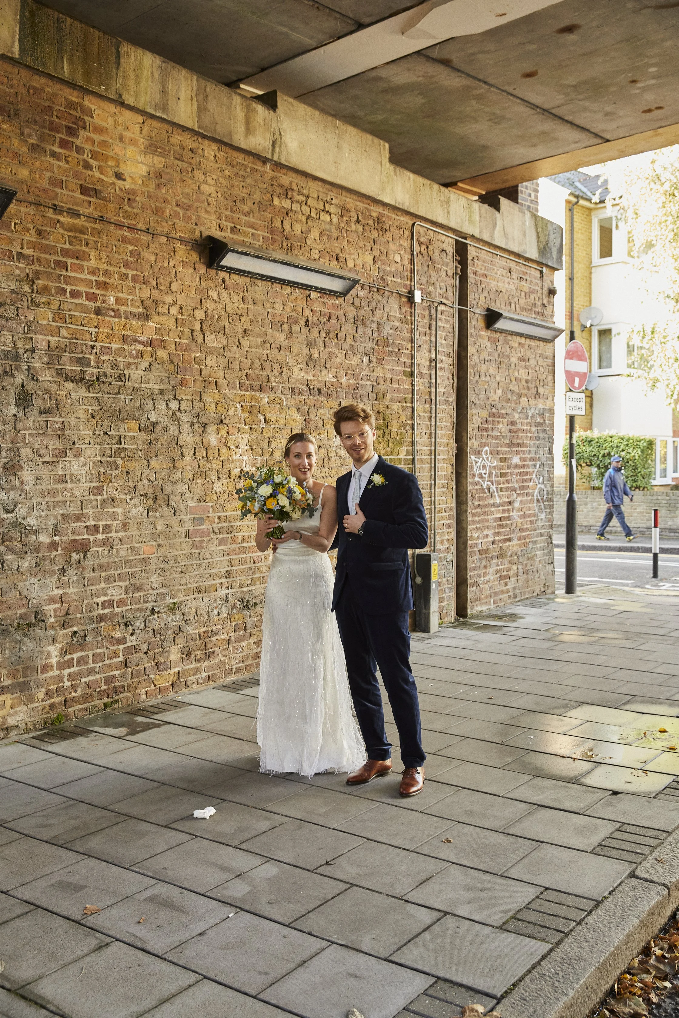 A bride and groom standing together outdoors against a brick wall, smiling. The bride is holding a bouquet of flowers and wearing a white wedding dress, while the groom is dressed in a dark suit and brown shoes.