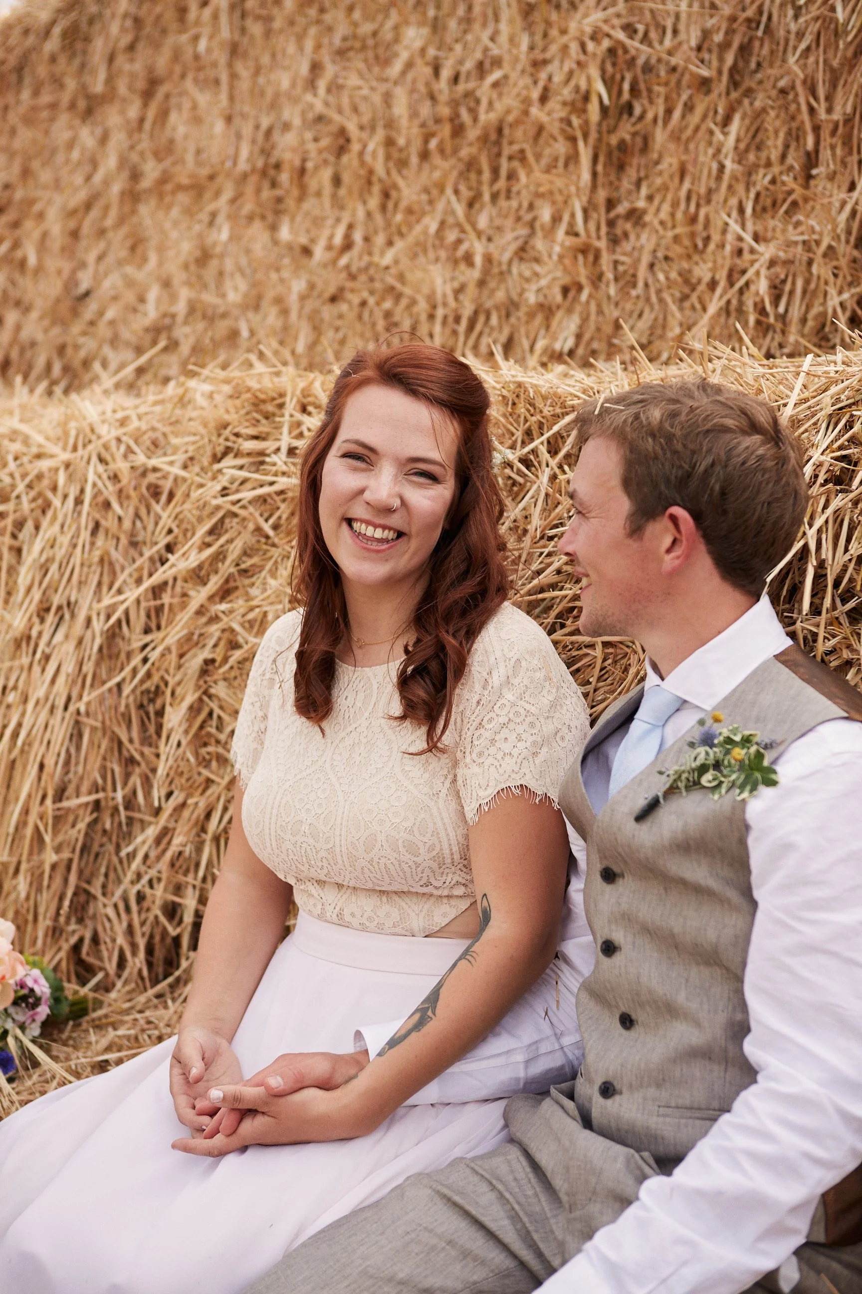 A smiling woman with long red hair and a tattoo on her arm sitting next to a man in a gray vest and white shirt, outdoors against a background of stacked hay bales, during a rustic wedding event.
