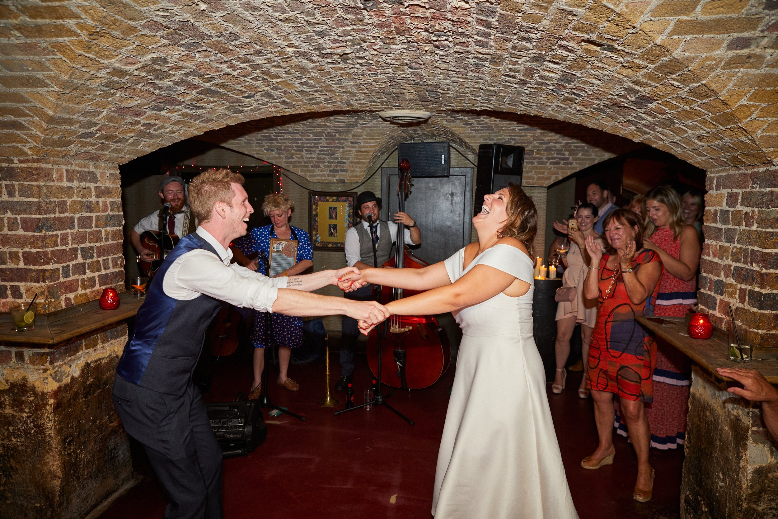 A bride and groom dancing and laughing at their wedding reception, surrounded by guests and a live band in a brick-walled underground venue.