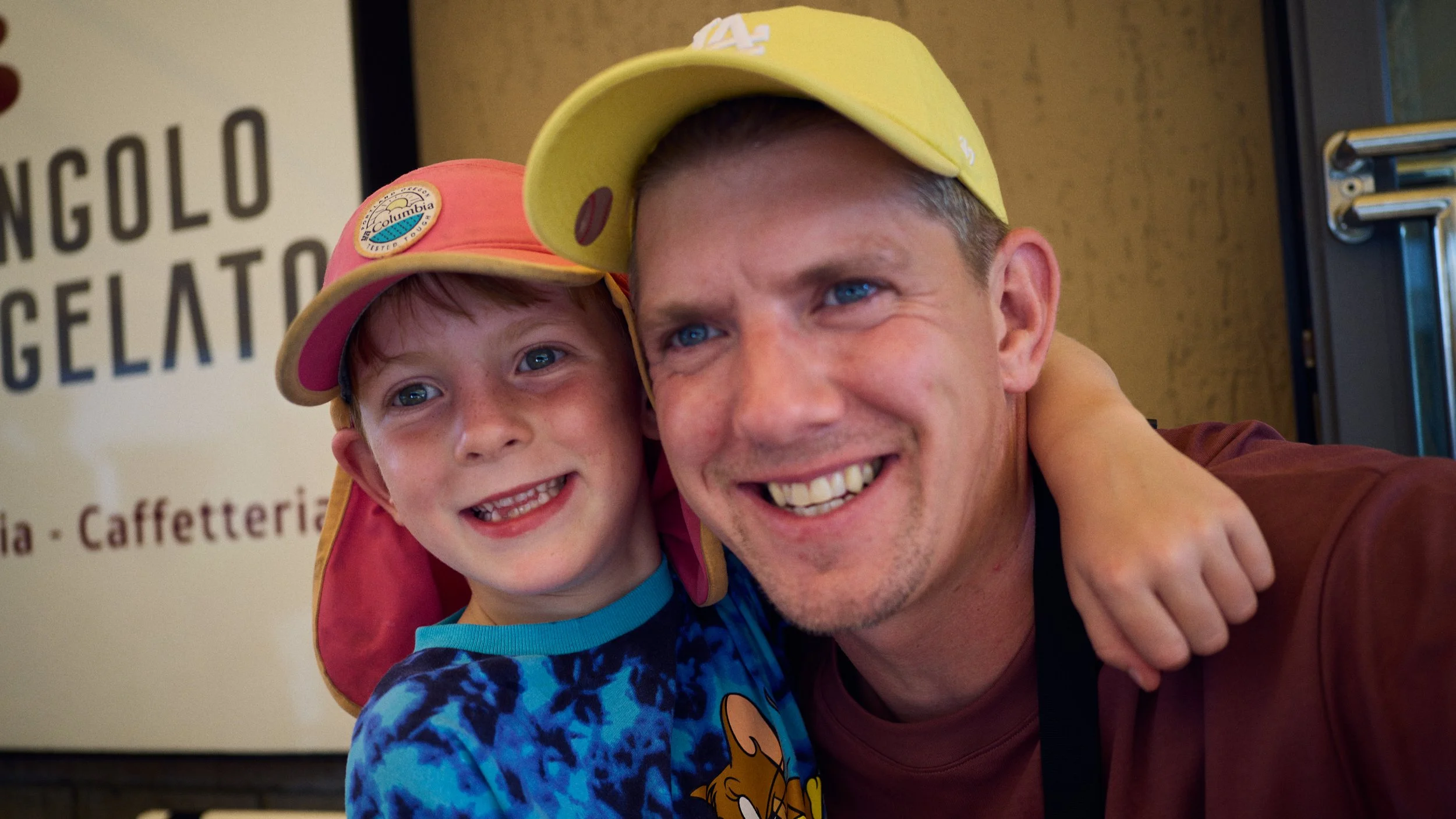 A smiling man and boy taking a close-up selfie. The boy is wearing a pink and yellow cap and a blue patterned shirt, while the man is wearing a yellow cap and a maroon shirt. They are inside a cafe or ice cream shop, with part of a sign in the background.